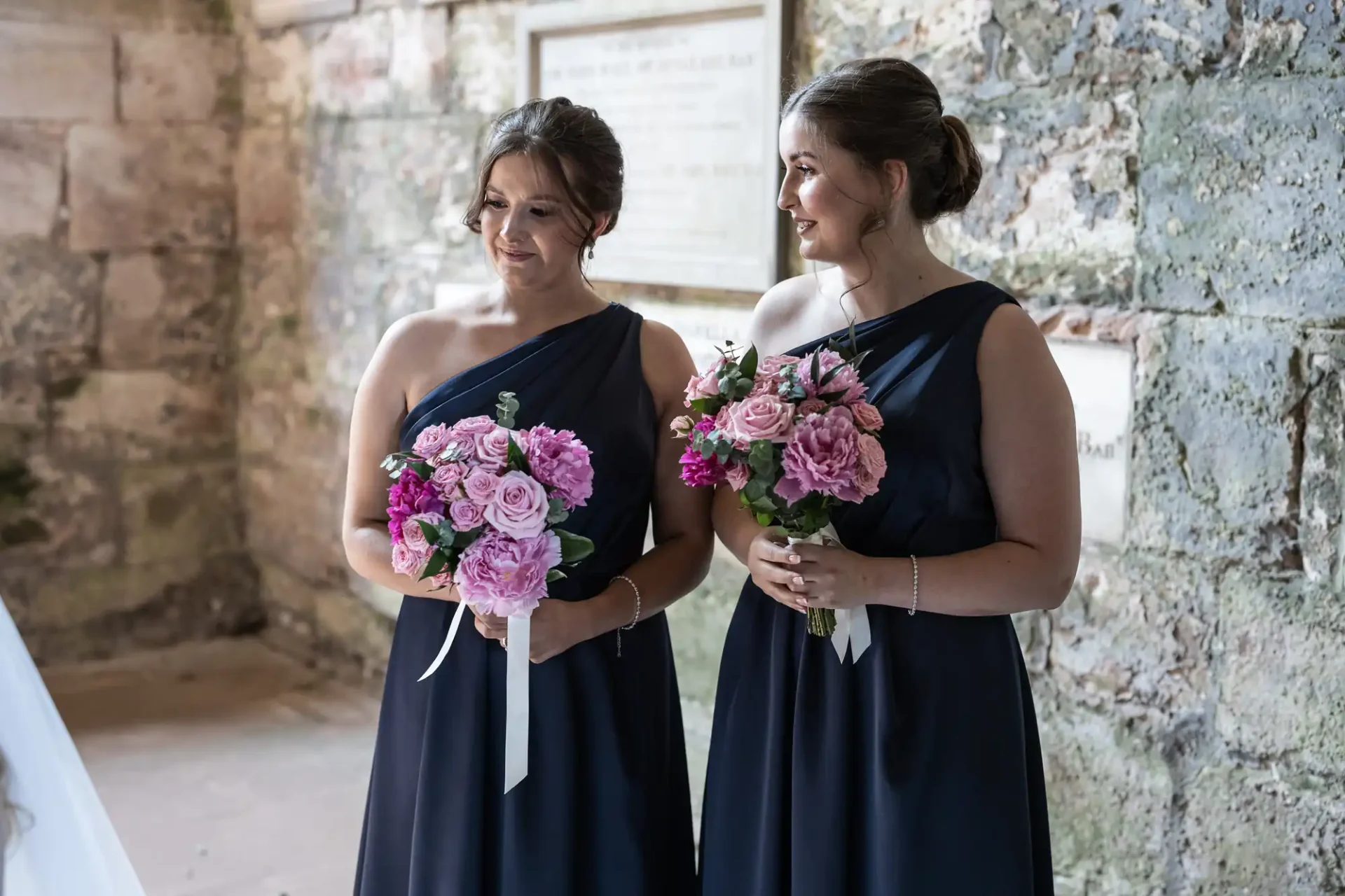 Two bridesmaids in navy dresses holding pink floral bouquets, standing by a rustic brick wall inside a venue.