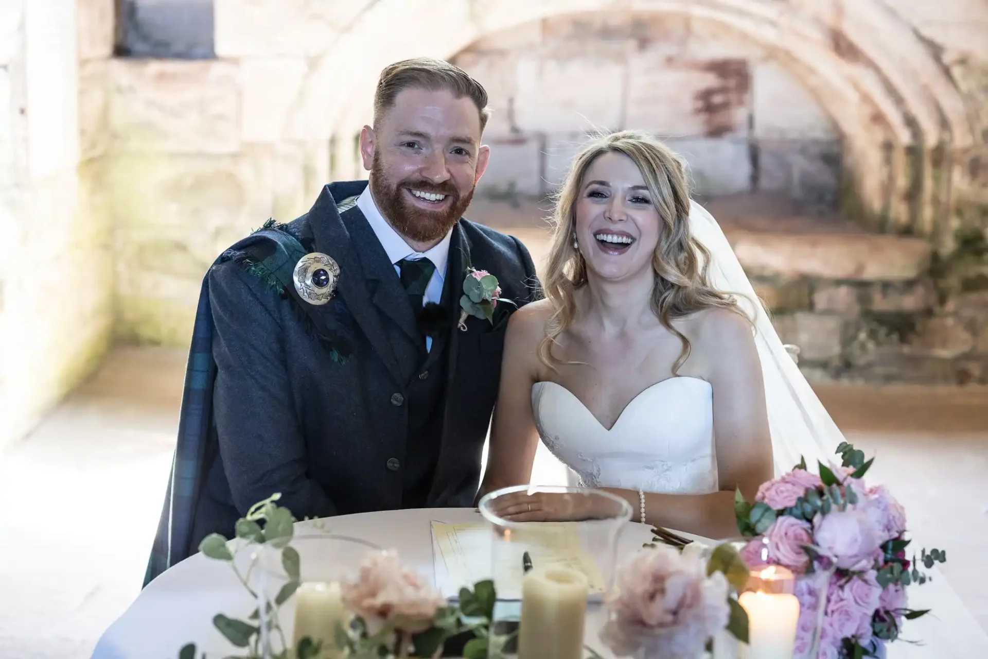 A joyful bride and groom sit at a wedding table, smiling broadly, in an elegant historic venue with stone arches in the background.