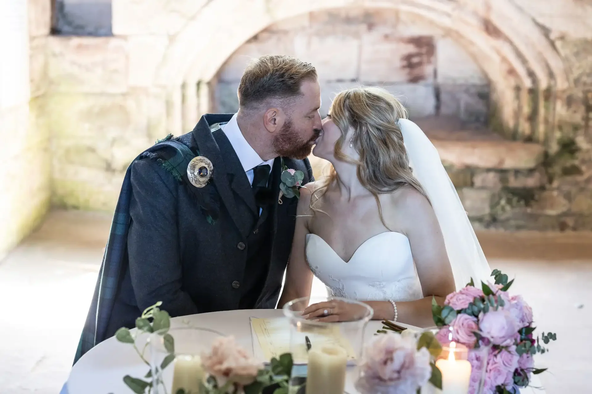 A bride and groom kiss across a table decorated with candles and flowers in a stone-arched venue.