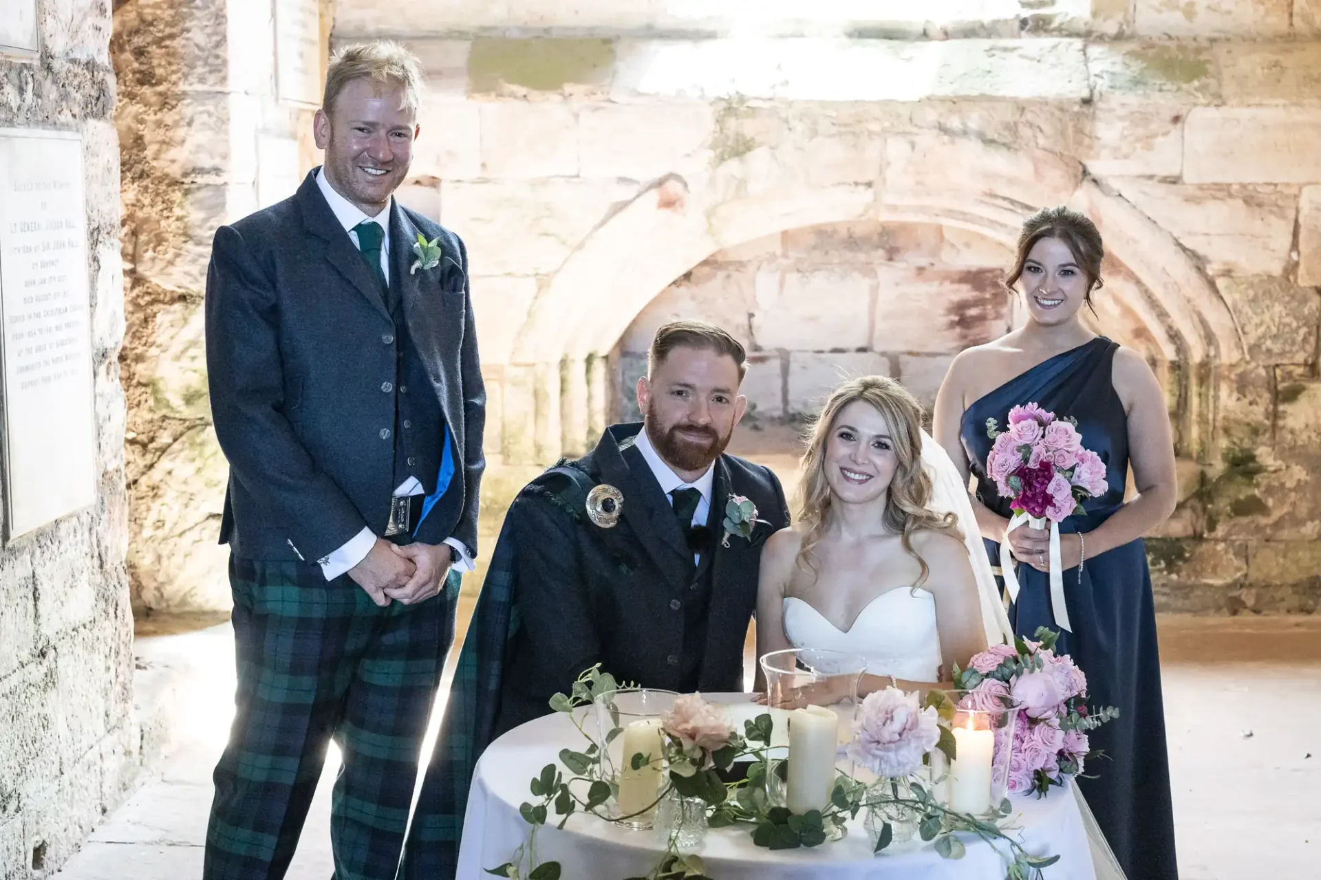 A bride and groom seated at a wedding table, smiling, flanked by a best man and a bridesmaid, in a stone-walled venue.