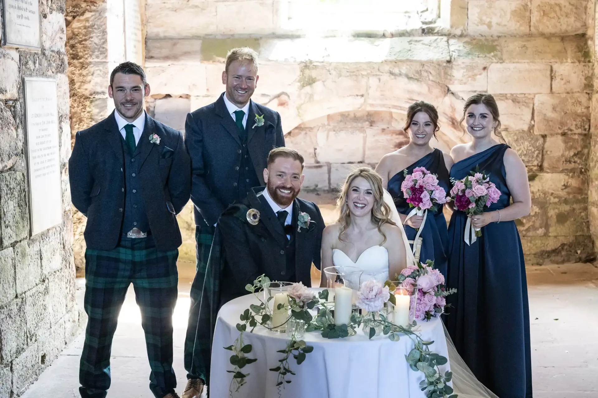 Three men and three women dressed elegantly, with two in tartan kilts, smiling around a table decorated with flowers inside a stone building.