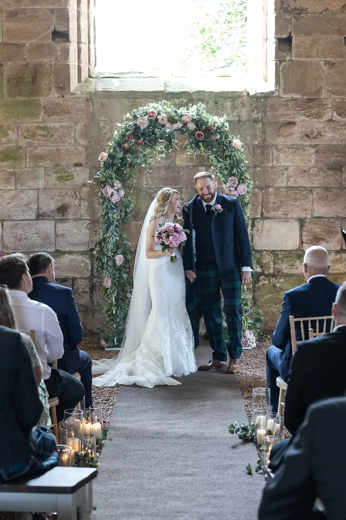 Bride and groom walking hand in hand down the aisle in a historic building, with guests seated on either side and a floral arch at the entrance.