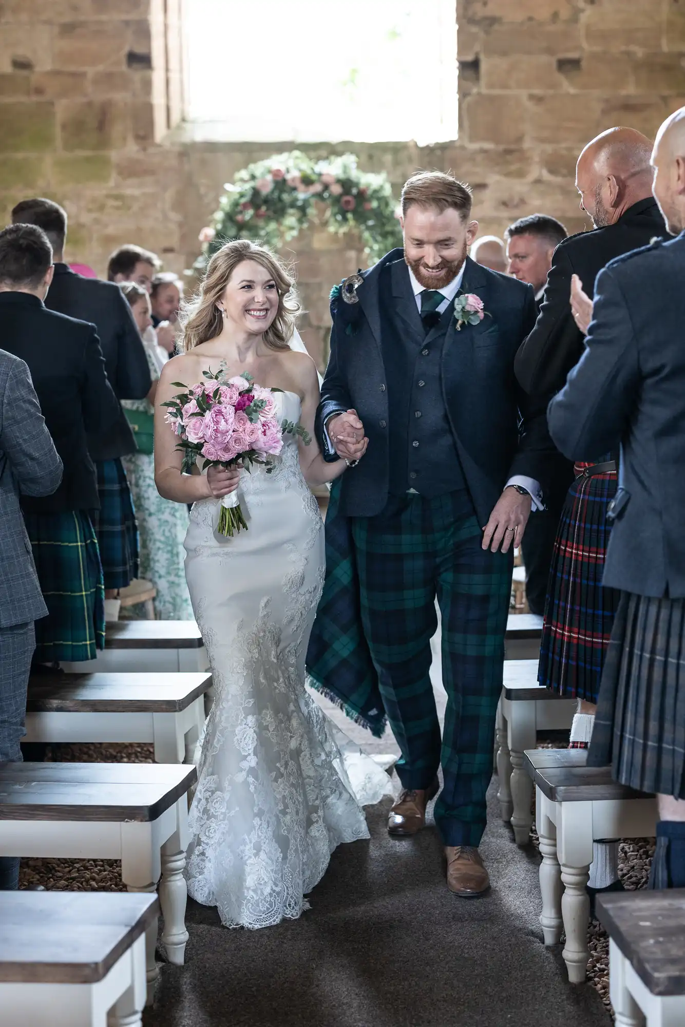 A bride and groom walk down the aisle, smiling, surrounded by guests; the groom wears a tartan kilt and the bride holds a bouquet.
