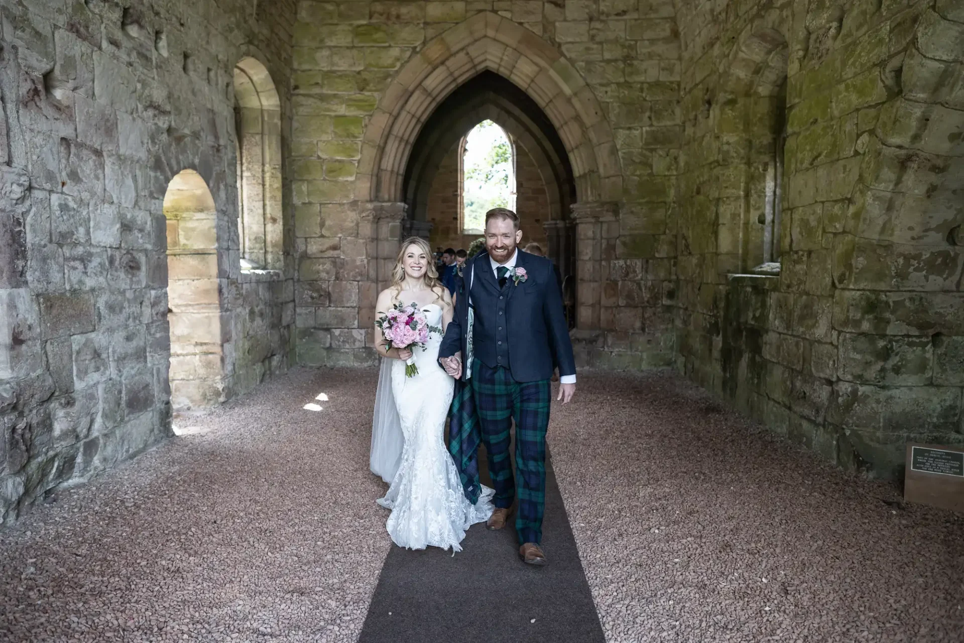 Bride and groom walking hand in hand through a stone archway at a historic site, the groom wearing a tartan kilt.