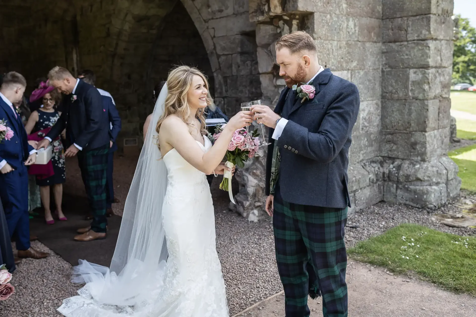 Bride in white dress and groom in kilt exchanging rings under an old stone arch, with guests in background.