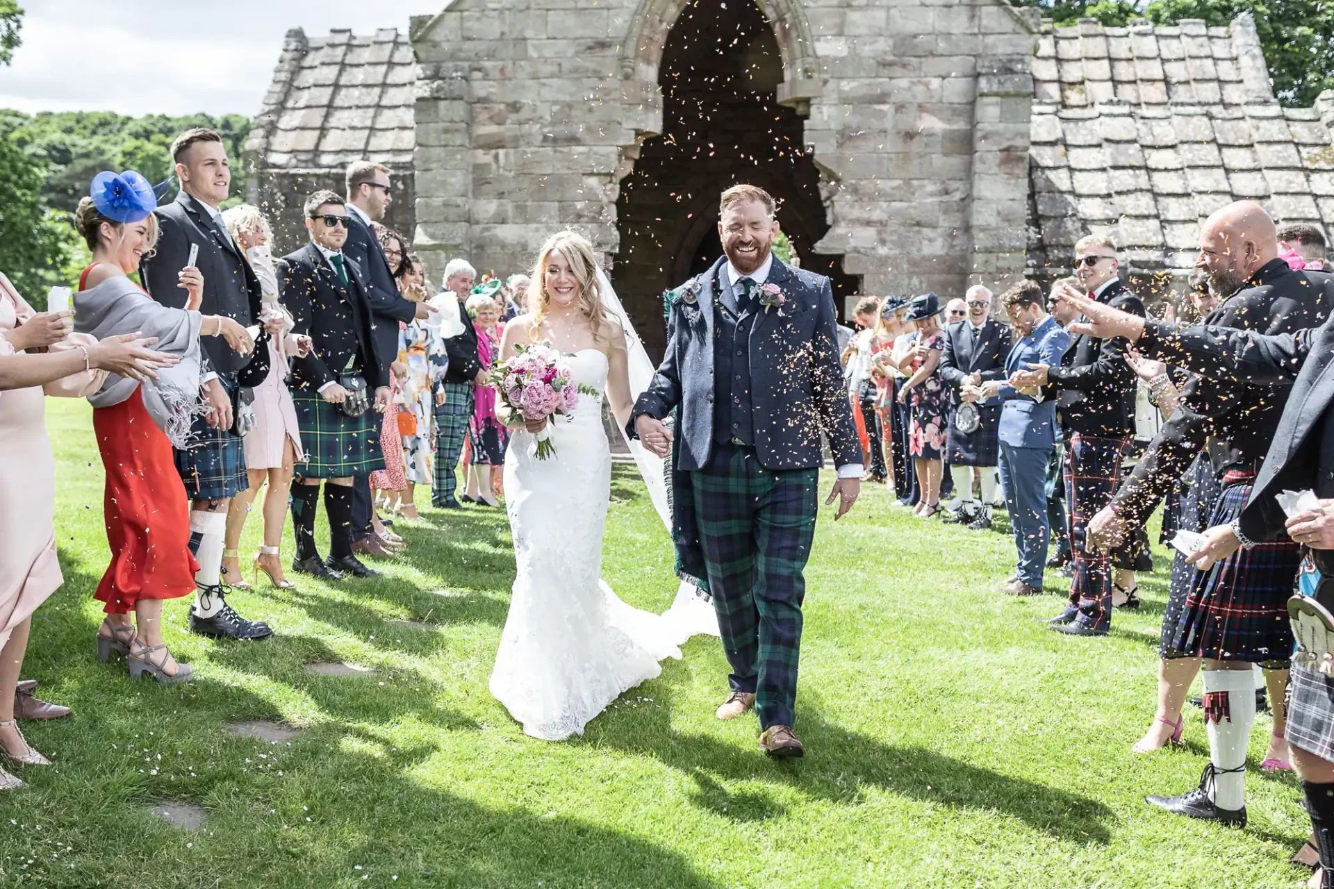 Bride and groom smiling and walking hand-in-hand while guests throw confetti at a sunny outdoor wedding ceremony.