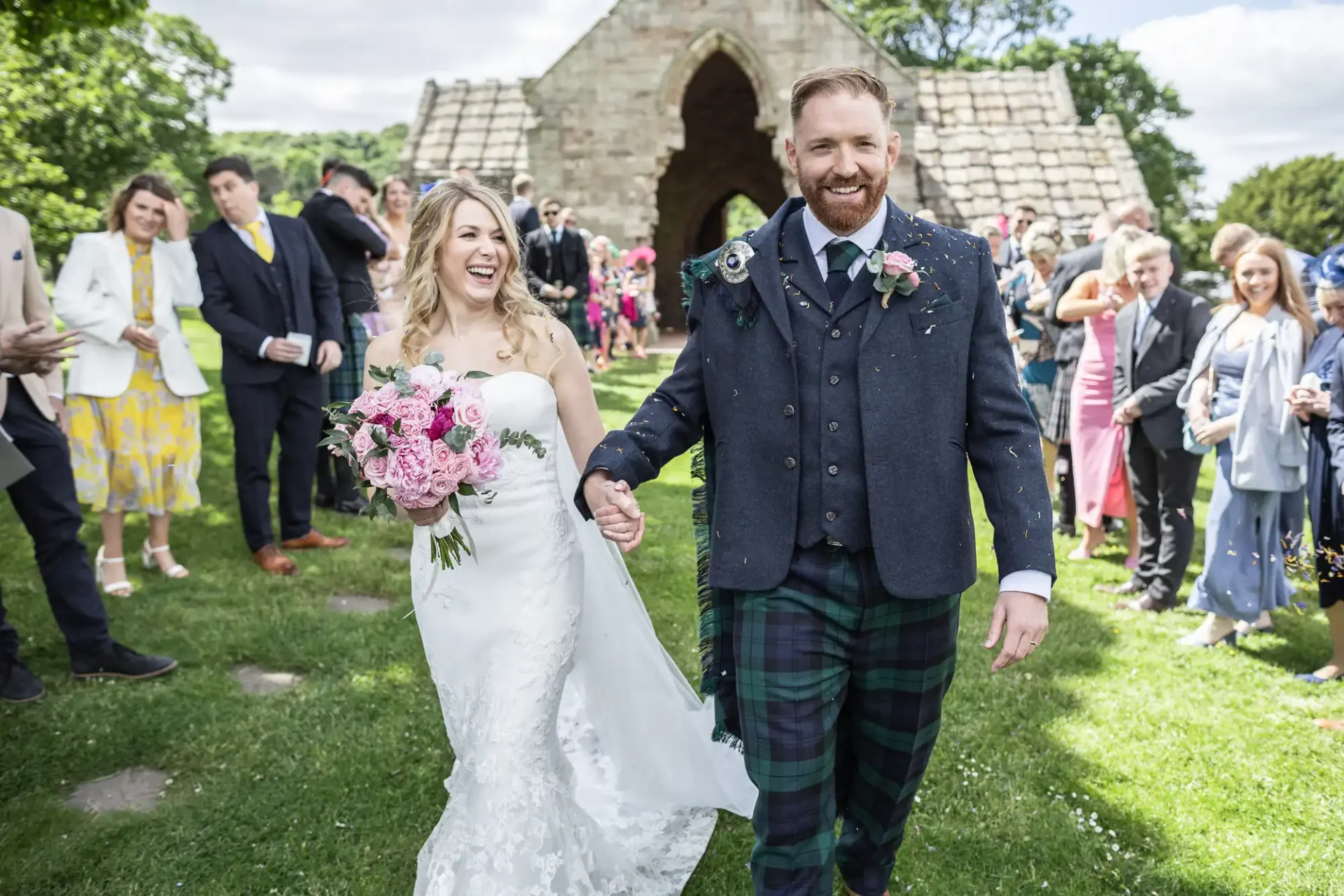 A bride and groom smiling and holding hands as they walk through guests after their wedding ceremony, the groom in a kilt and the bride in a white dress.