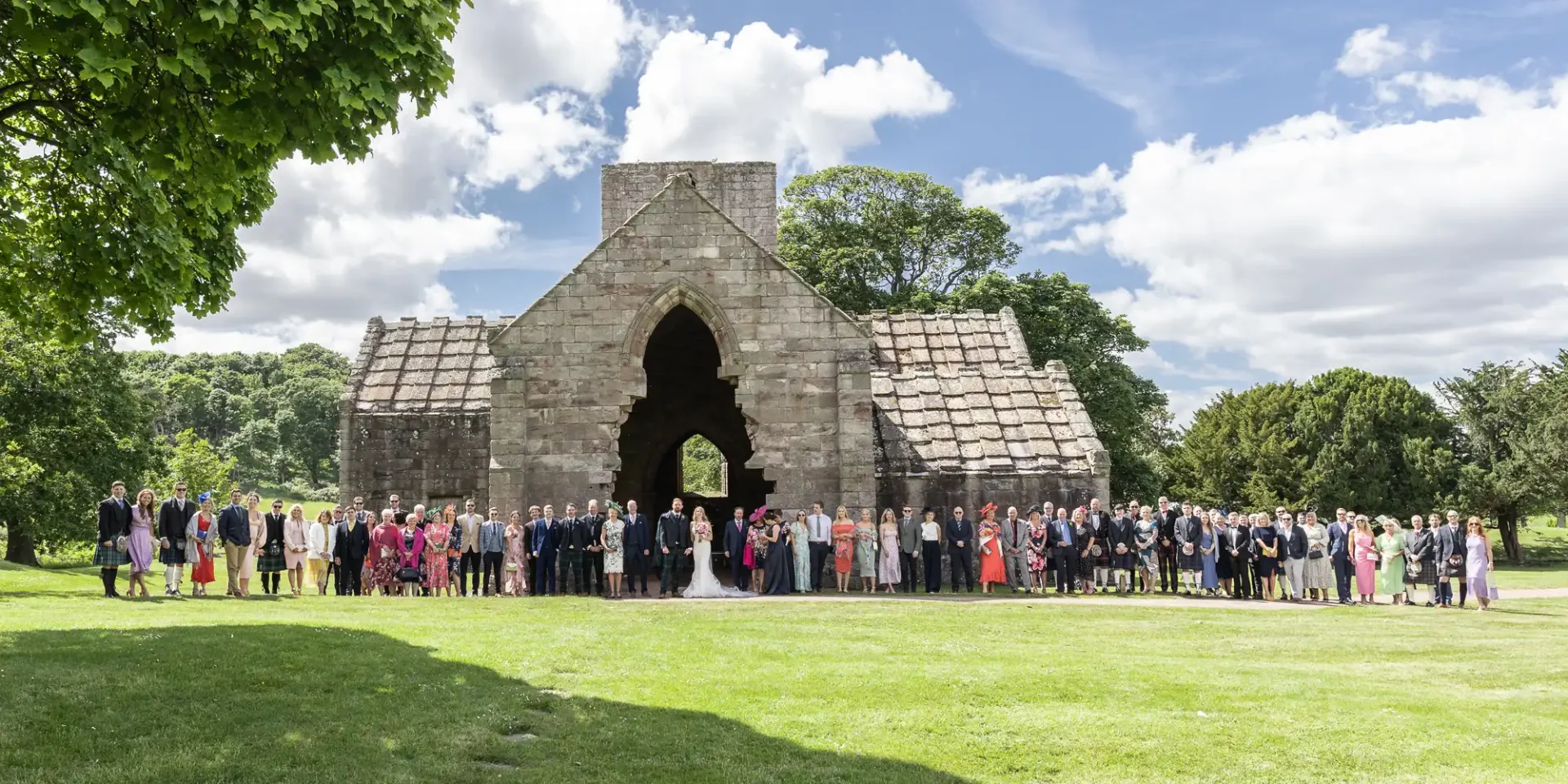 Large group of people gathered for a wedding ceremony in front of an old stone archway in a lush green park.
