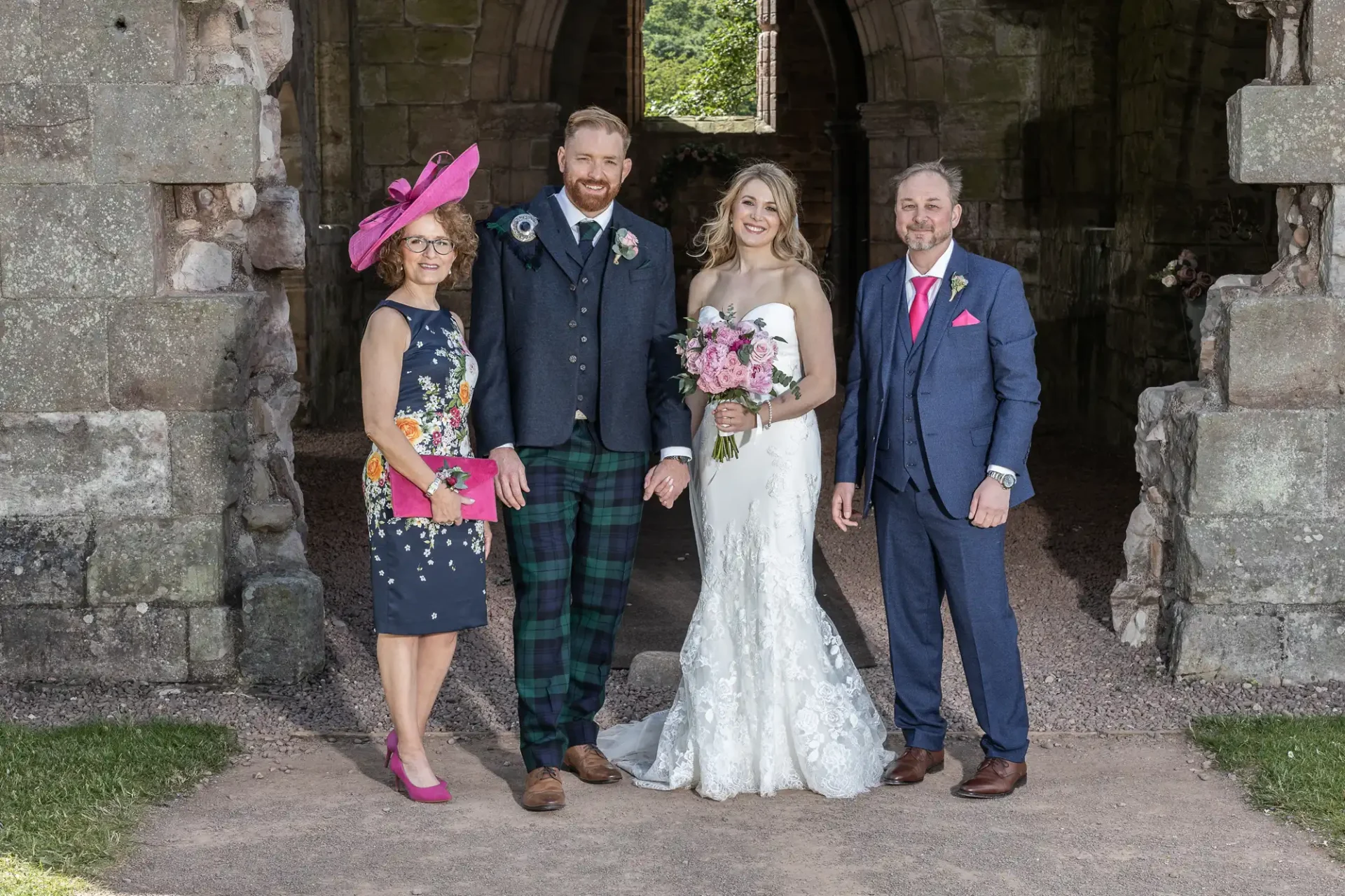 A wedding photo of a smiling bride and groom with two guests, men in suits and a woman in a pink hat, standing in a historic stone archway.