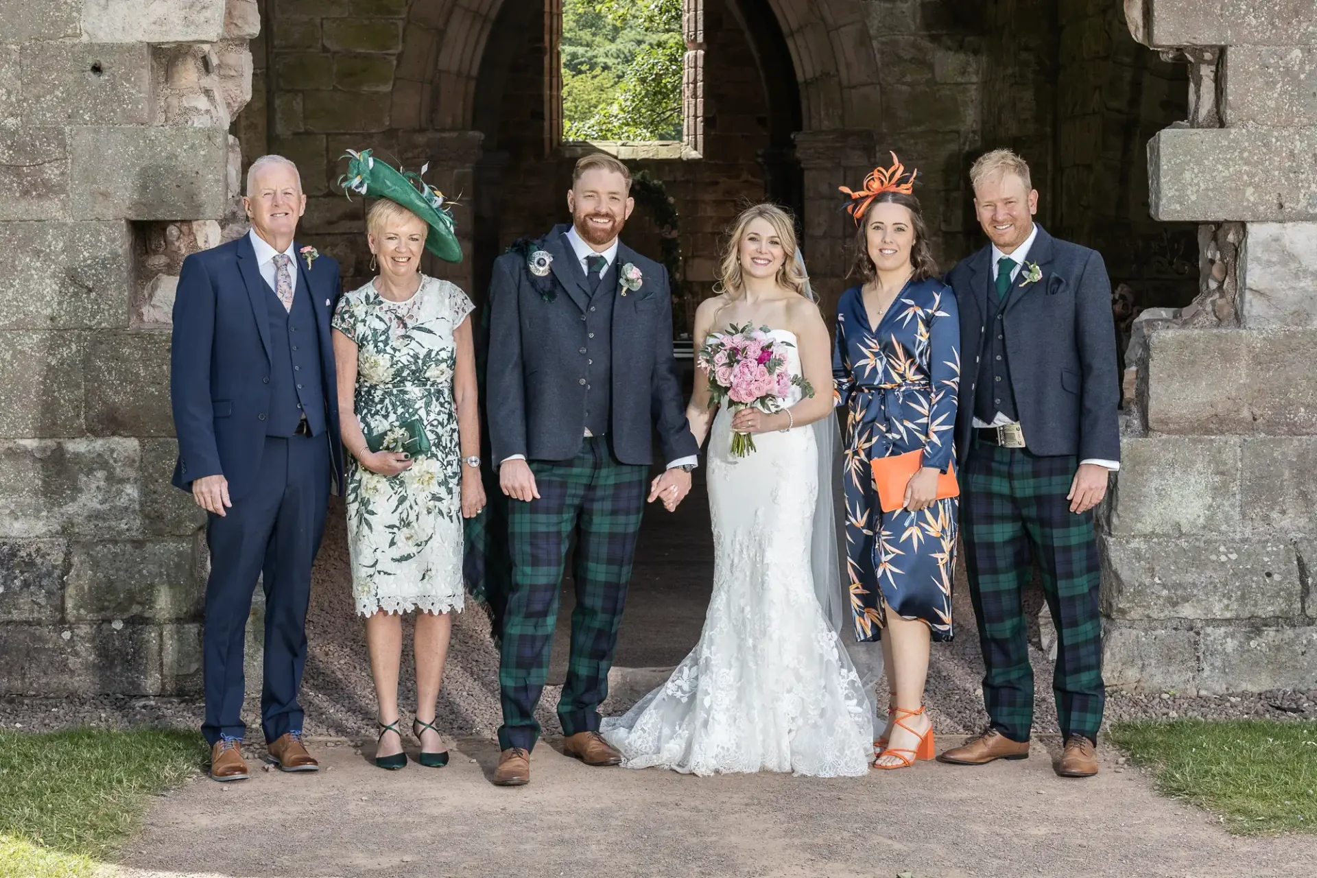 A wedding group photo featuring a newlywed couple with four guests, all smiling, in formal attire with tartan patterns, standing by stone ruins under a clear sky.