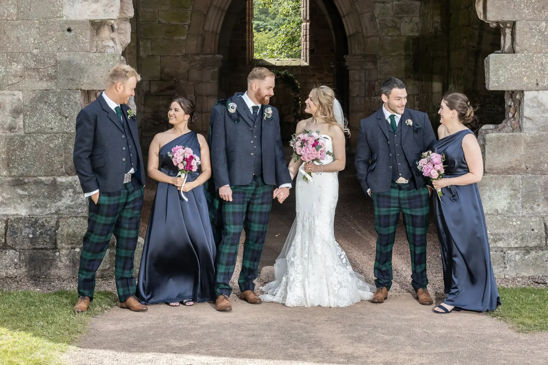 A wedding party of three couples, the men in tartan kilts and tweed jackets, and the women in blue dresses, posing in a stone archway.