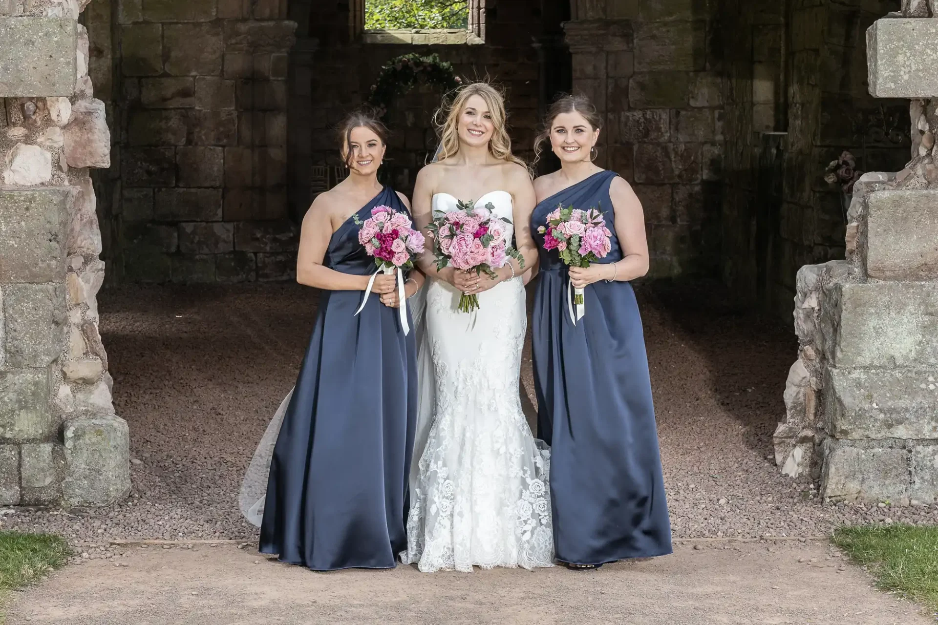 A bride in a white dress flanked by two bridesmaids in navy dresses, all holding pink bouquets, standing in a stone archway.