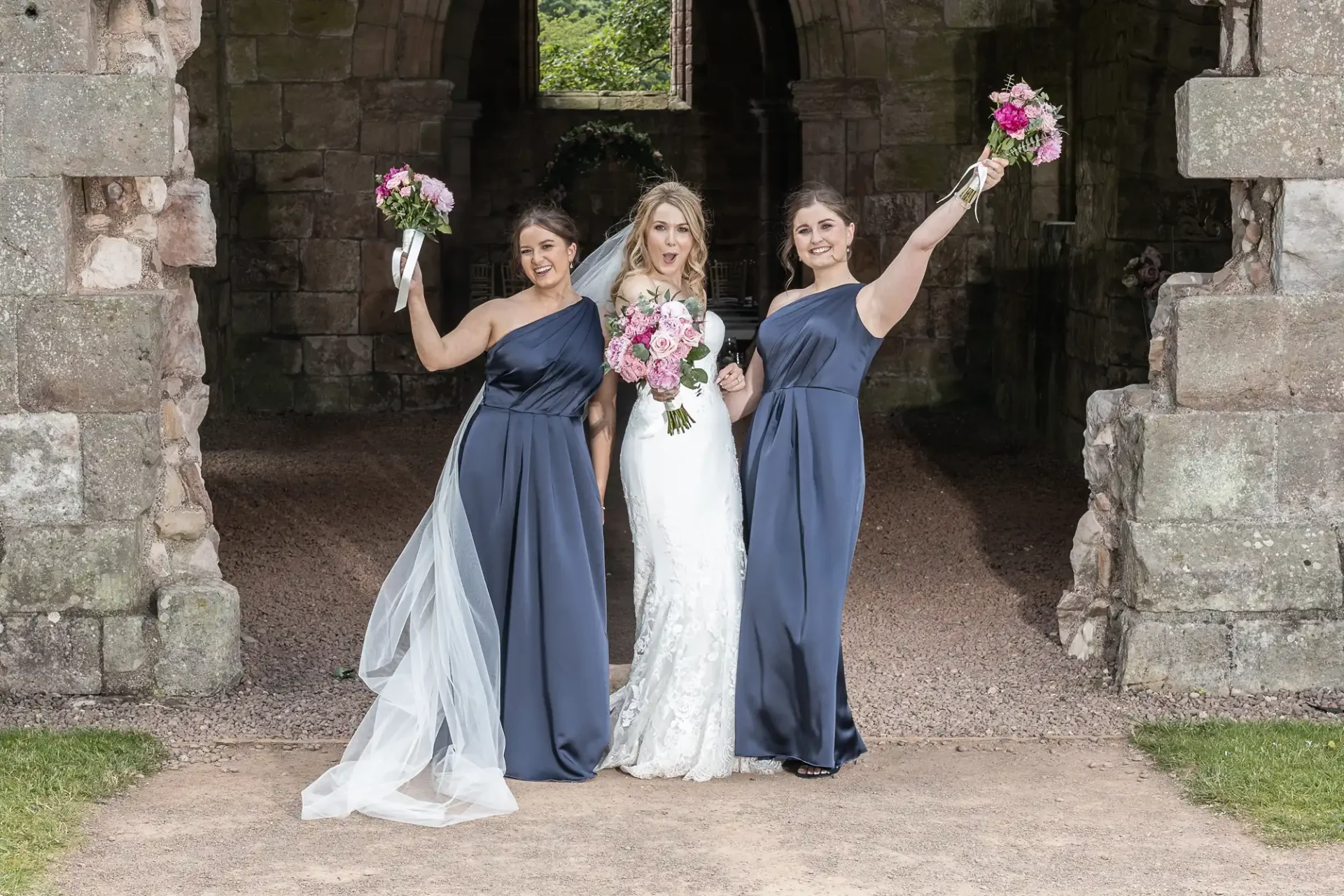 Three women in formal dresses, one in white and two in navy, holding bouquets, pose joyfully at a stone archway.