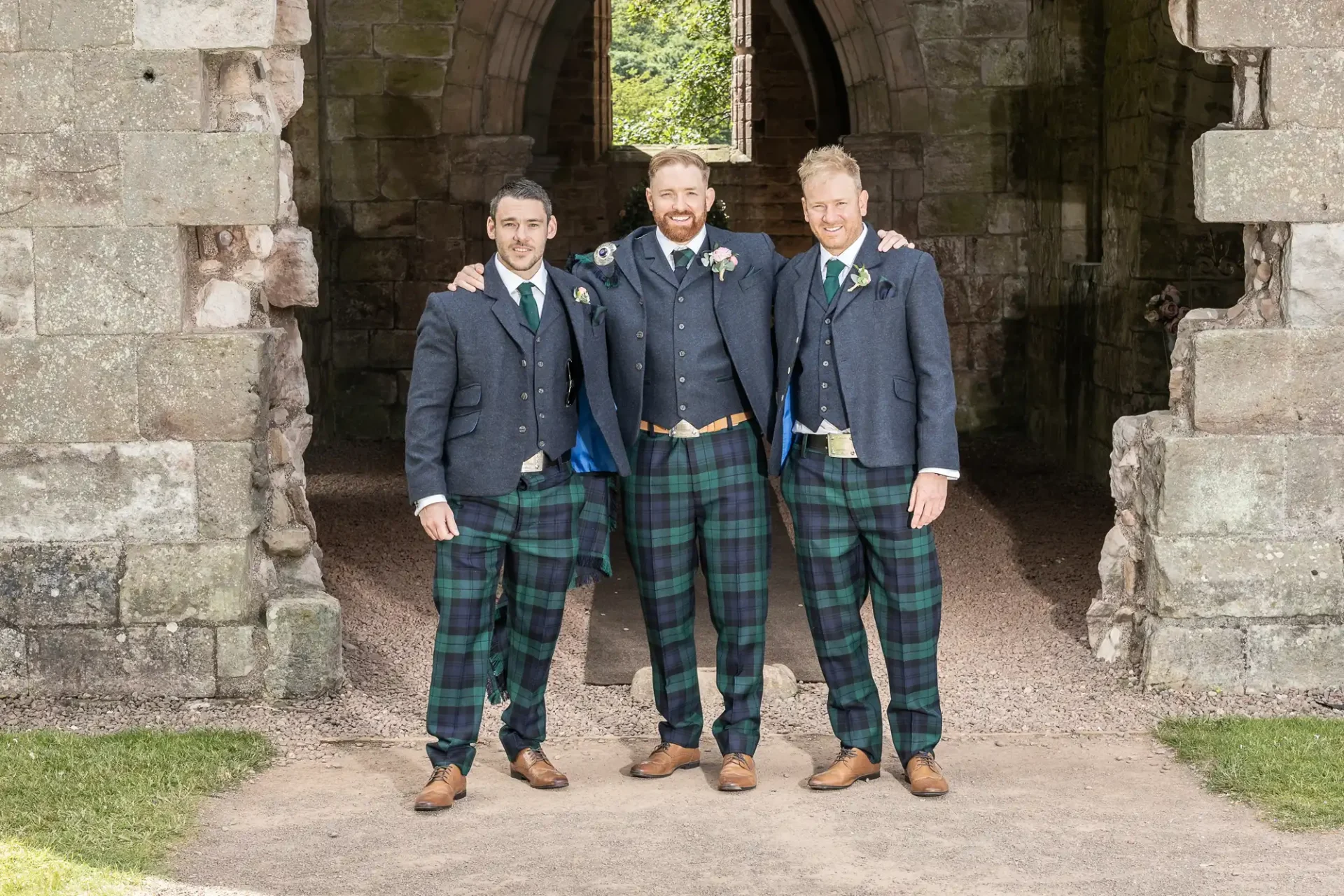 Three men in tartan kilts and tweed jackets posing with arms around each other's shoulders in front of an old stone archway.