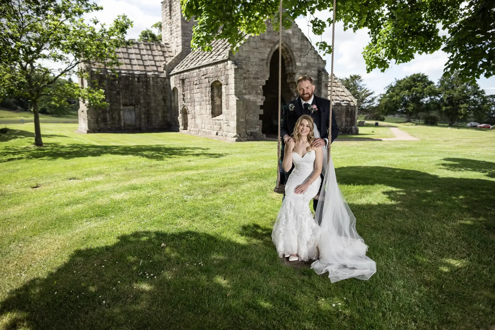A bride and groom smiling under a tree, near a historic stone church on a sunny day with a clear blue sky and lush green grass.