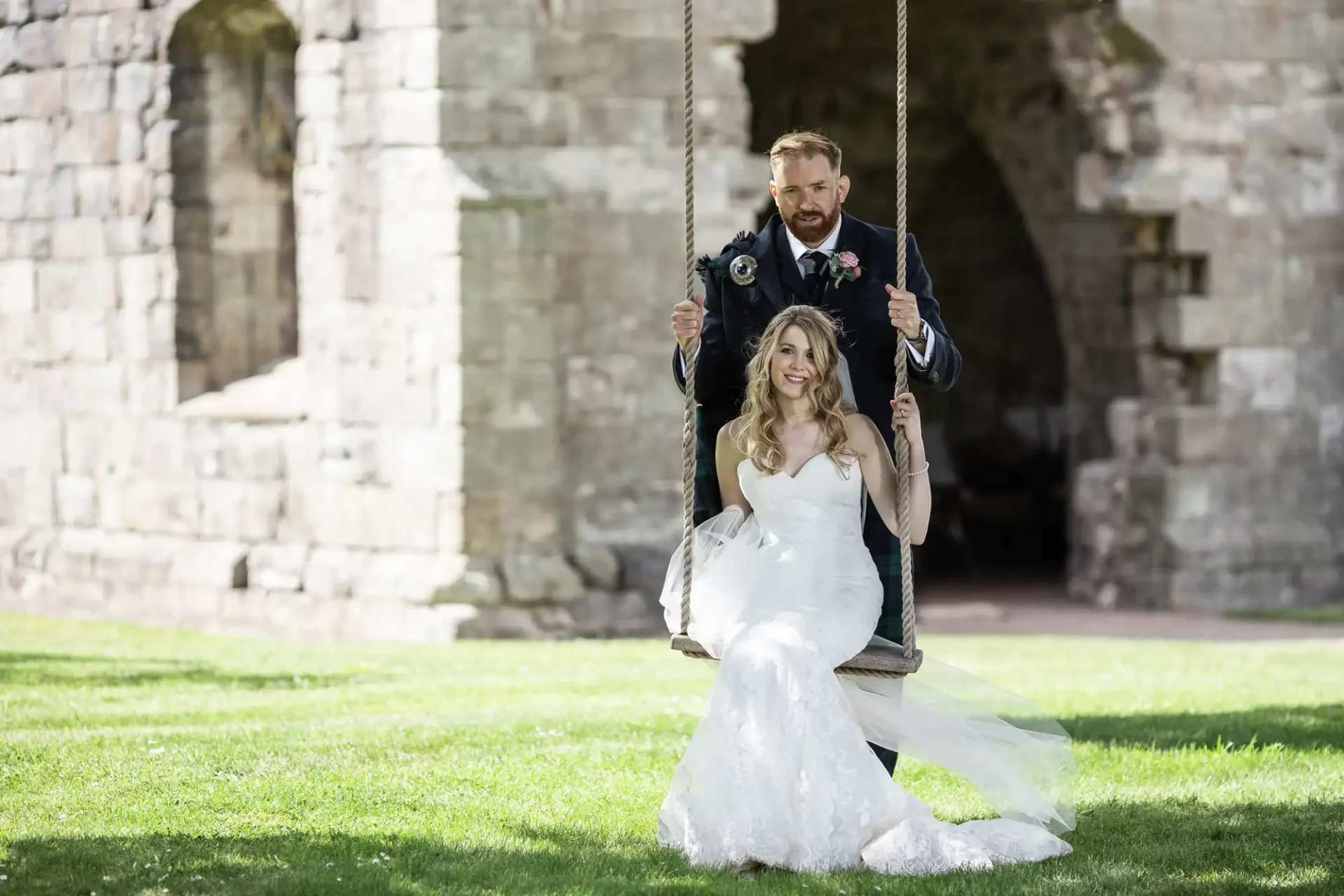 A bride and groom smiling on a swing, set against the backdrop of an old stone archway in a lush green park.