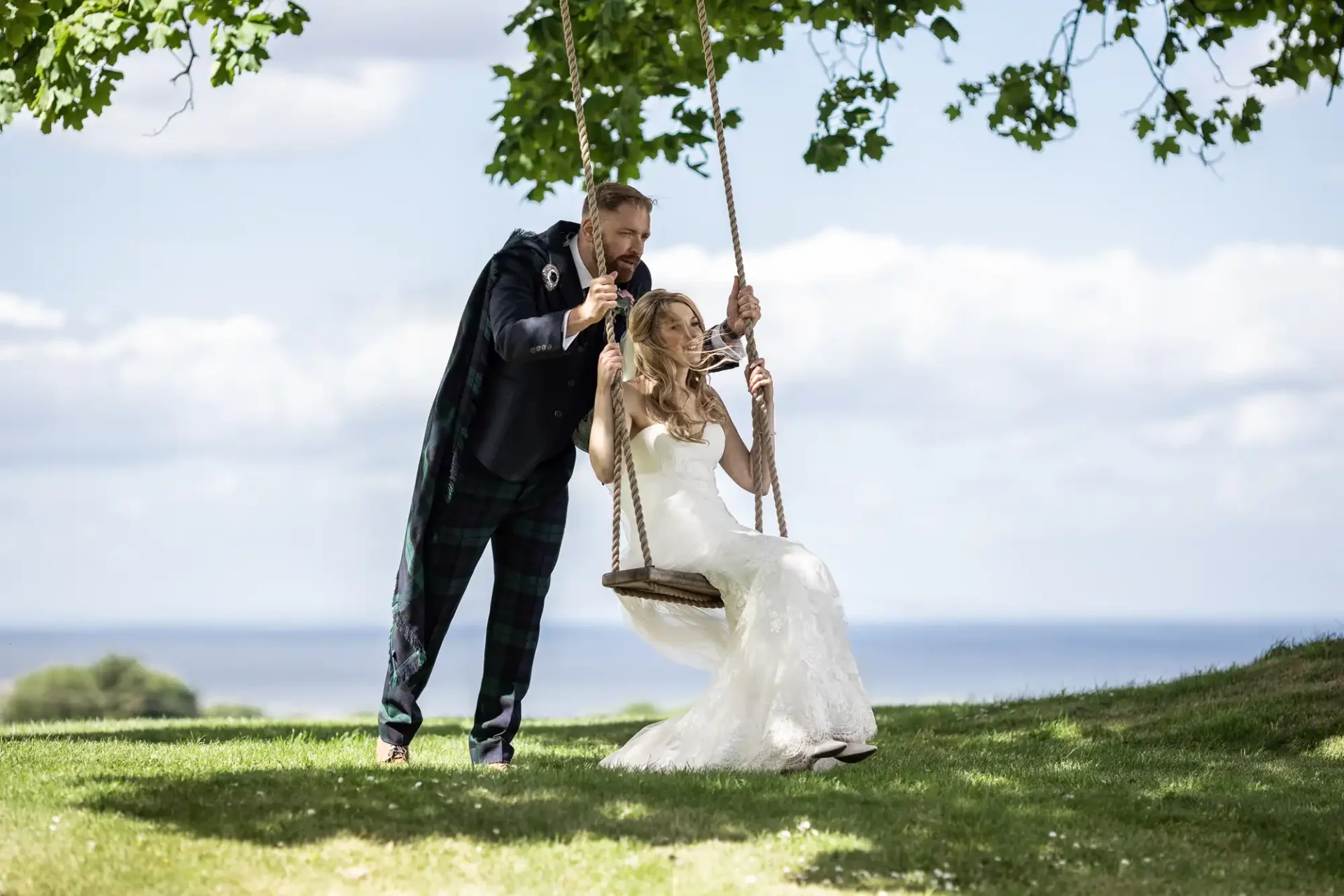 A groom in a tartan kilt pushing a bride on a swing under a tree, with a scenic lake and clear sky in the background.