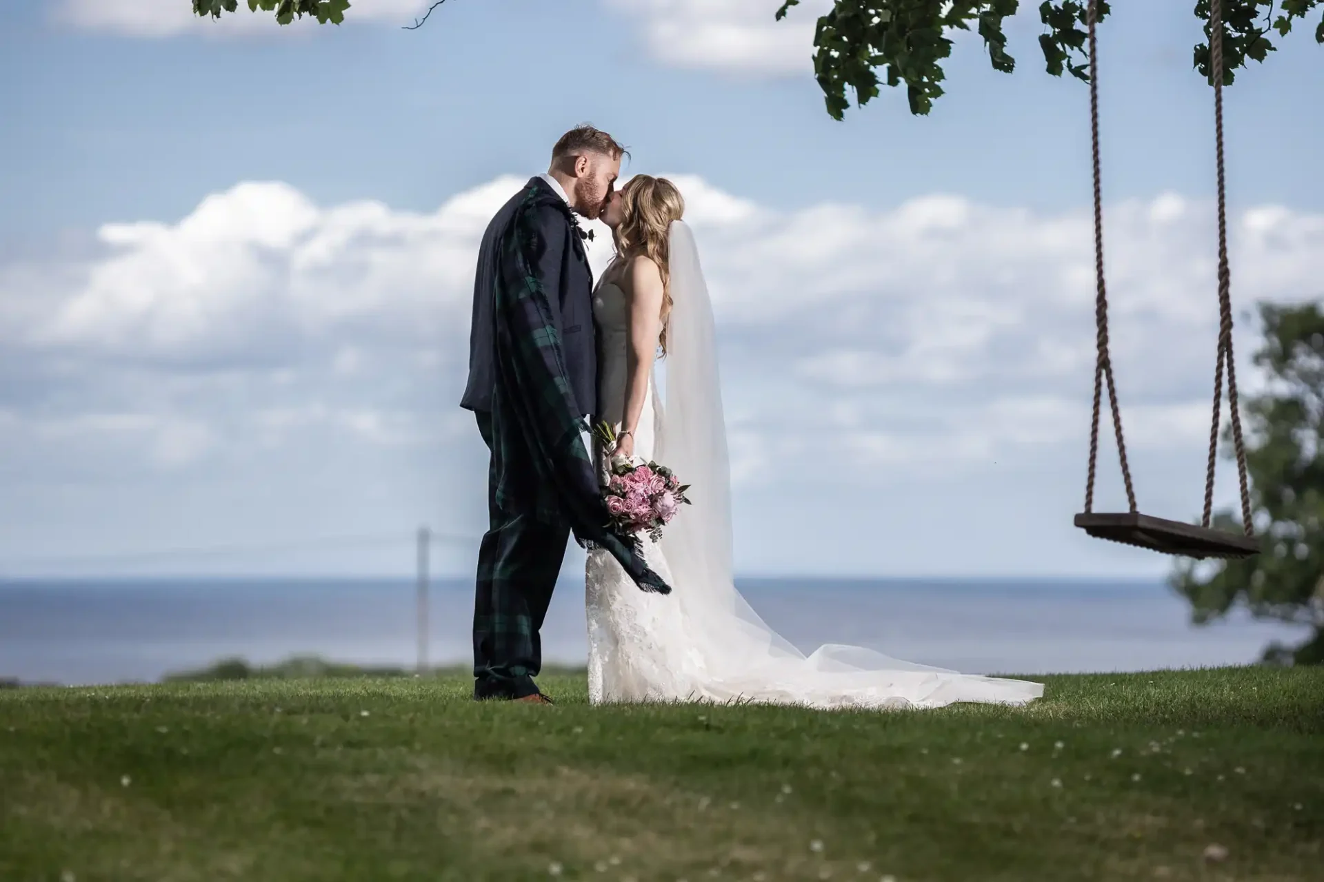 A bride and groom kissing outdoors on a sunny day, with a swing hanging from a tree and a sea backdrop.