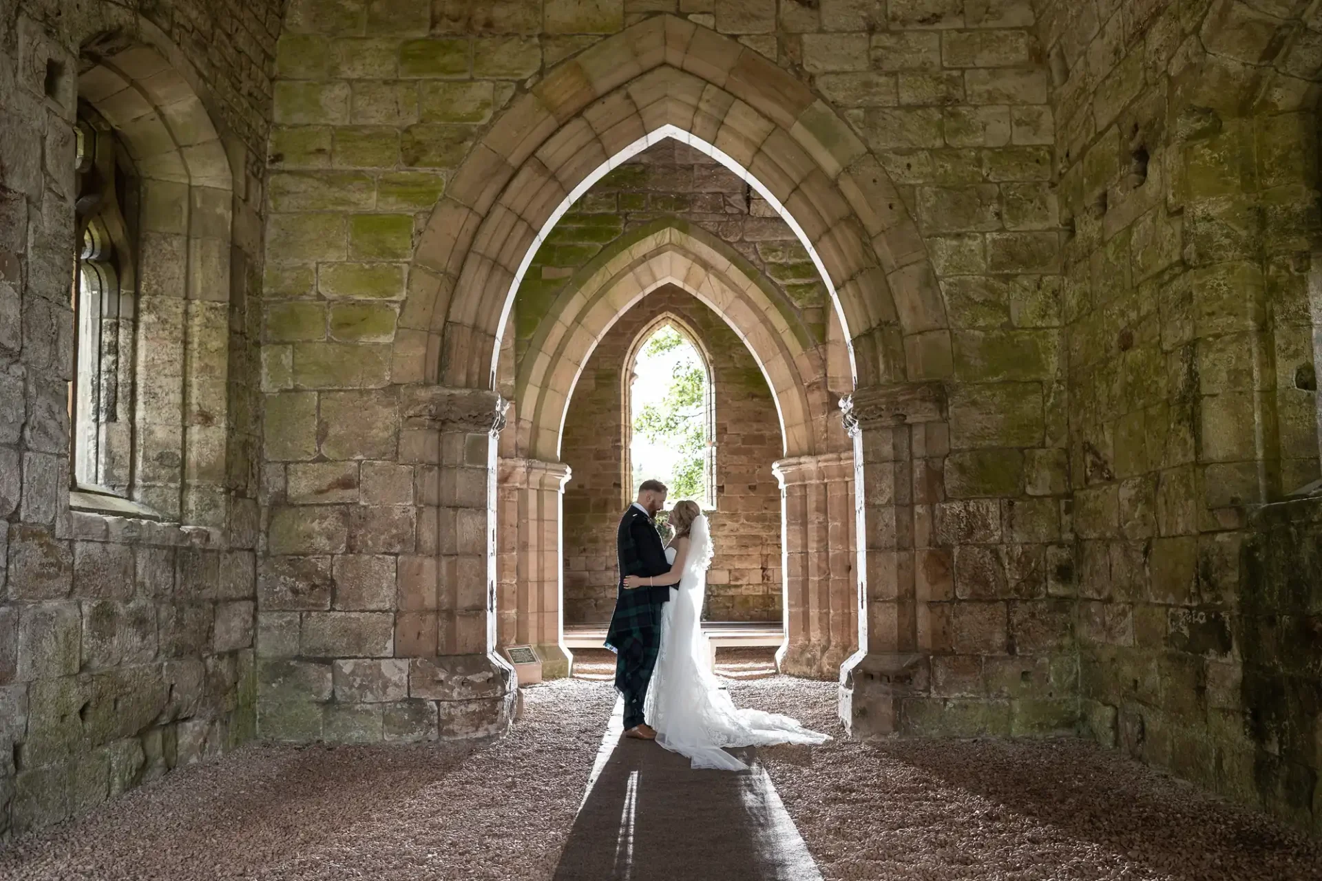 A bride and groom embrace under the arched doorways of an ancient stone building, with natural light illuminating them from behind.