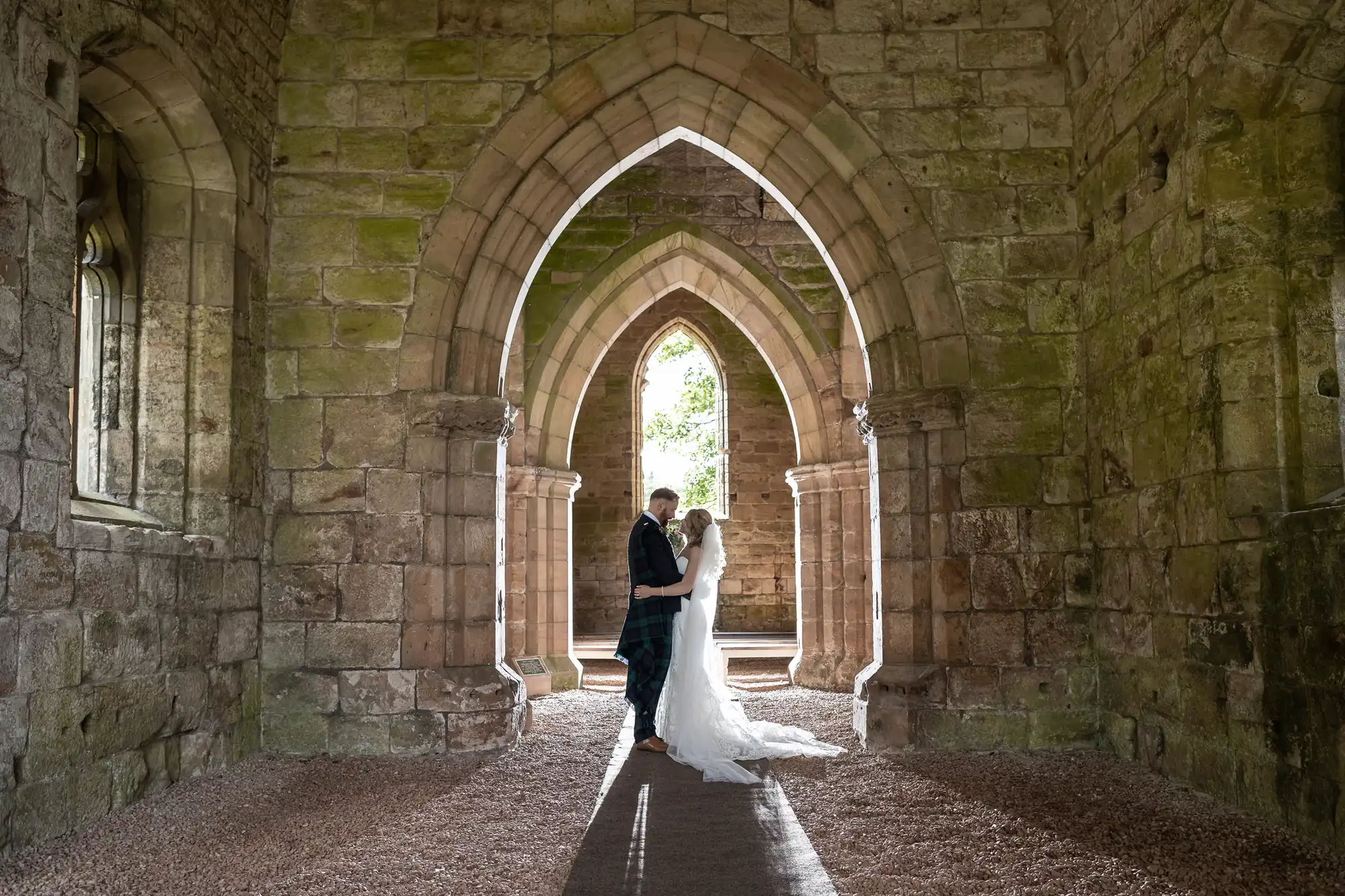 A bride and groom embrace under the arched doorways of an ancient stone building, with natural light illuminating them from behind.