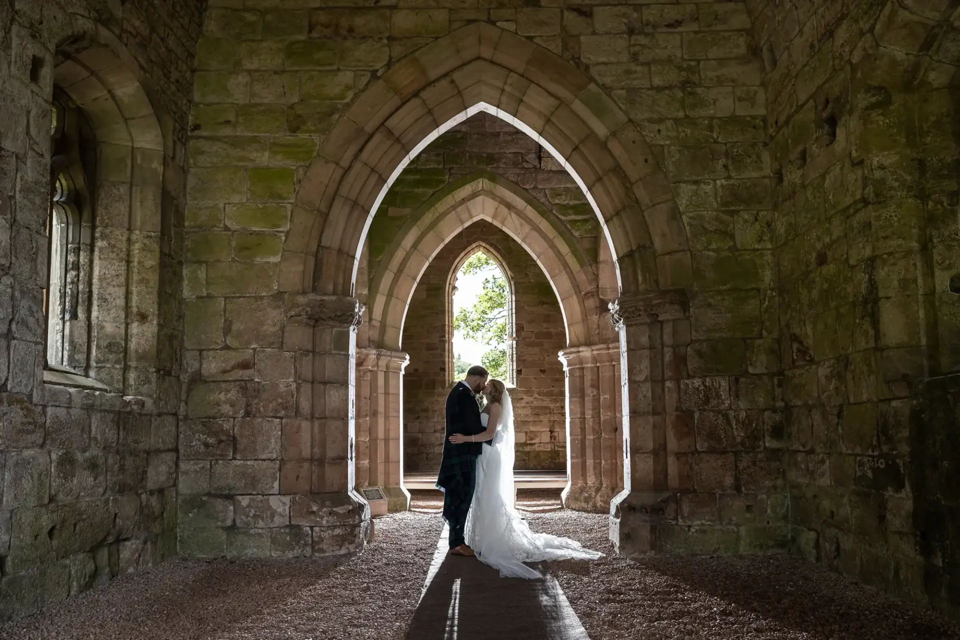 A bride and groom share an embrace under the arches of a stone church ruin, with trees visible in the background.