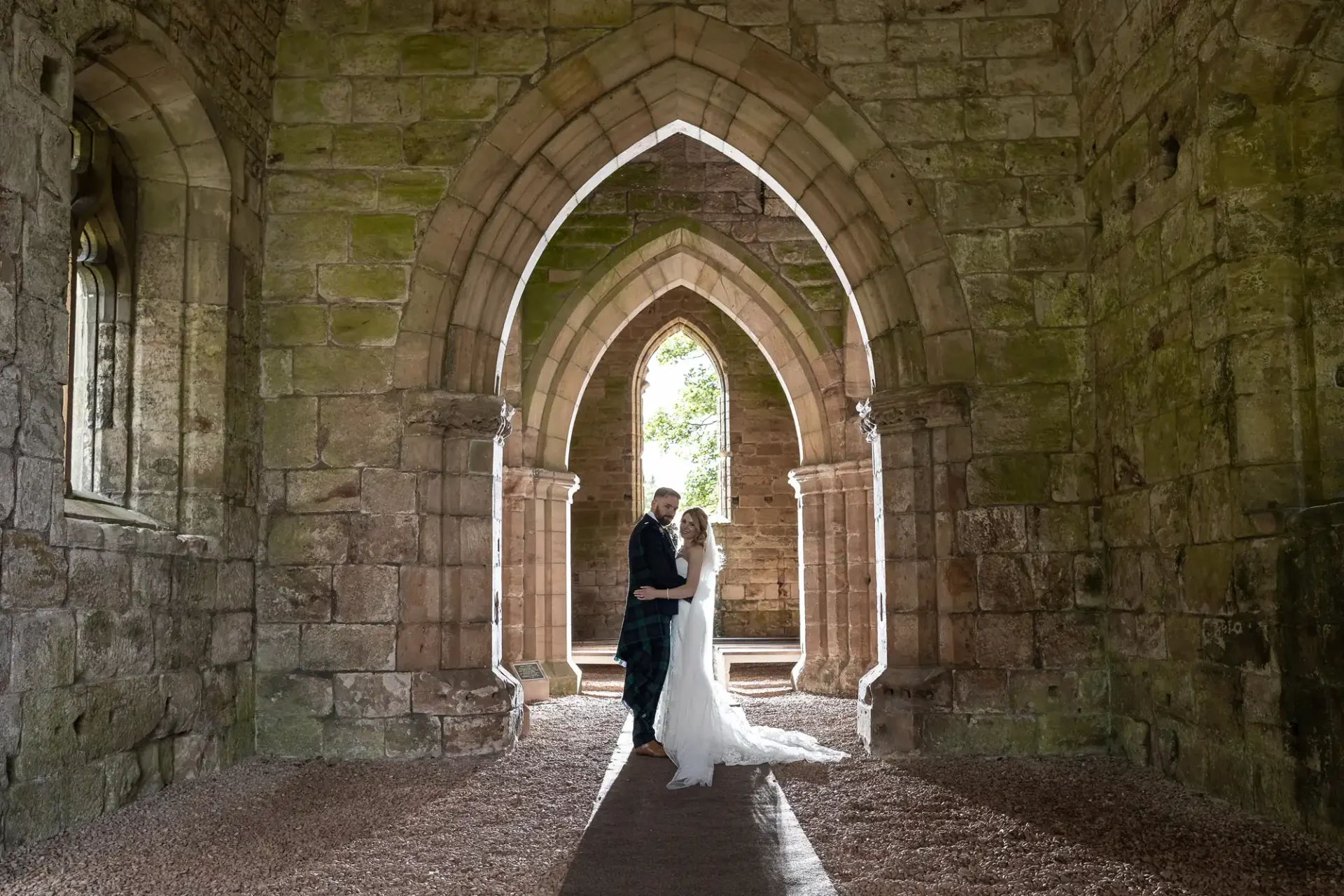 A couple embraces under the arches of an ancient stone ruin, with sunlight filtering through the windows.