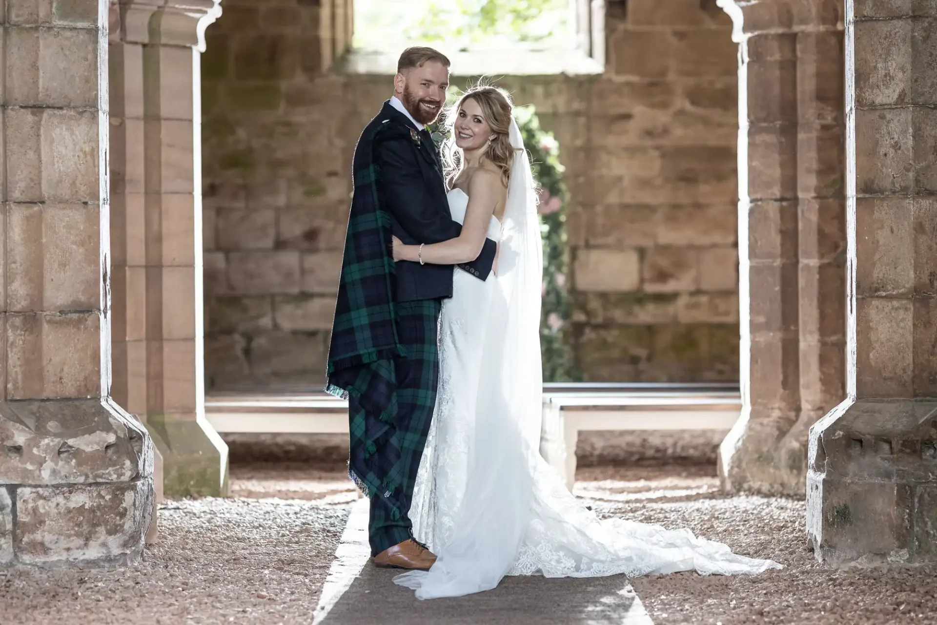 A bride and groom smile and embrace each other in a historic stone corridor, with the groom in a kilt and the bride in a long white dress.