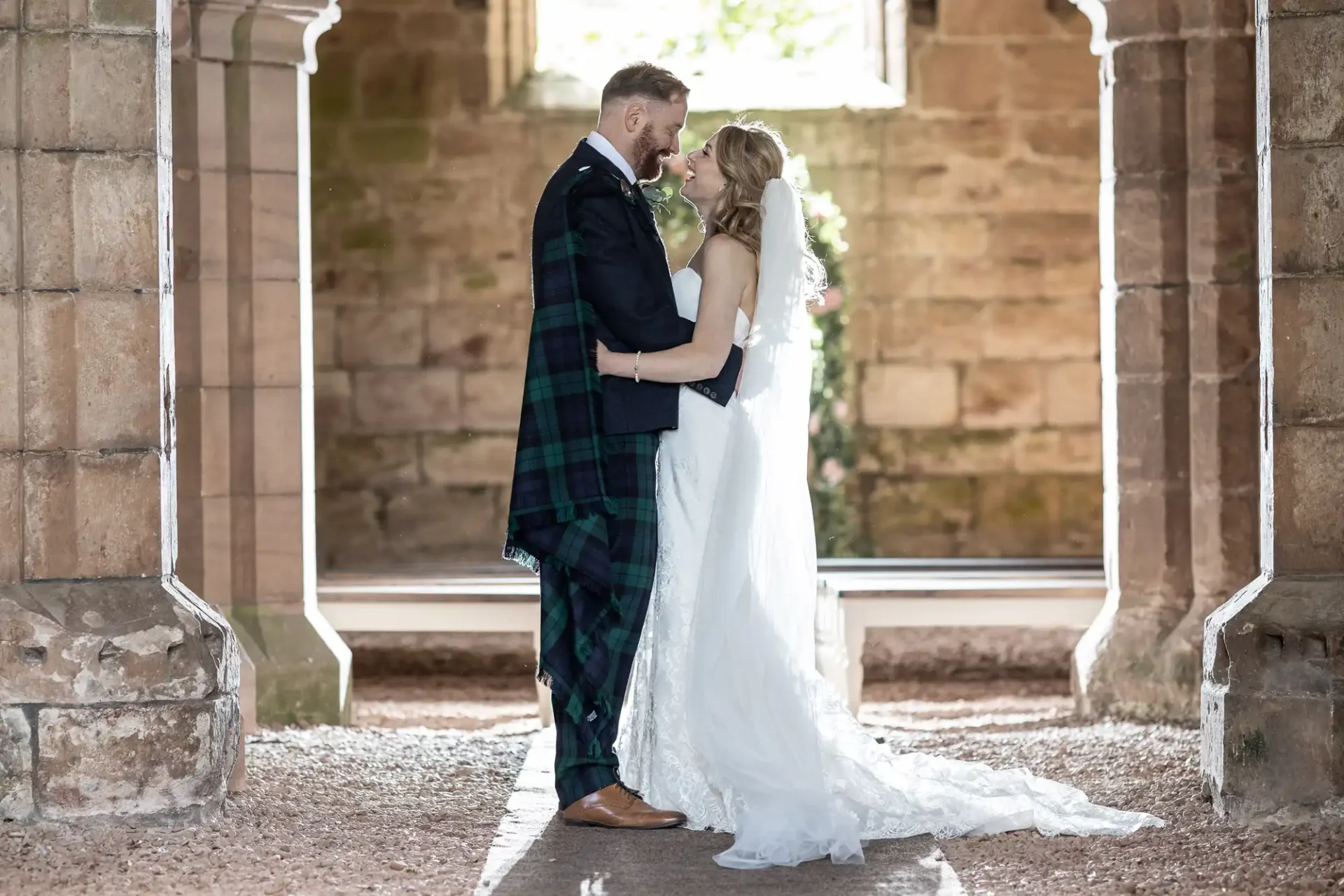 A bride and groom share a romantic moment in a sunlit stone archway, the groom in a tartan kilt and the bride in a white dress with a long train.