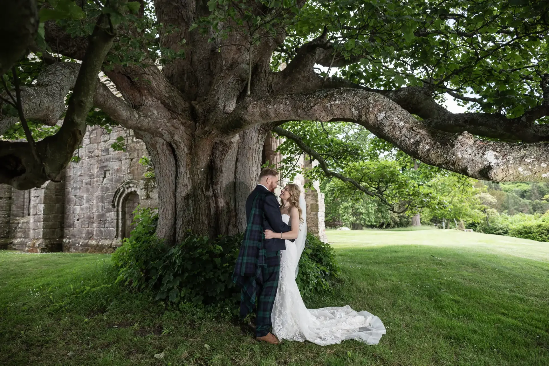 A bride and groom stand beneath an ancient tree, softly embracing, with an old stone building in the background.