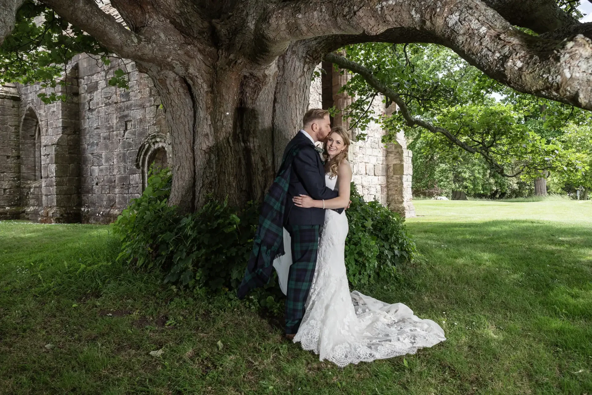 A bride and groom embracing under an ancient tree, with the groom wearing a tartan kilt and the bride in a lace gown, near old stone ruins in a lush green setting.