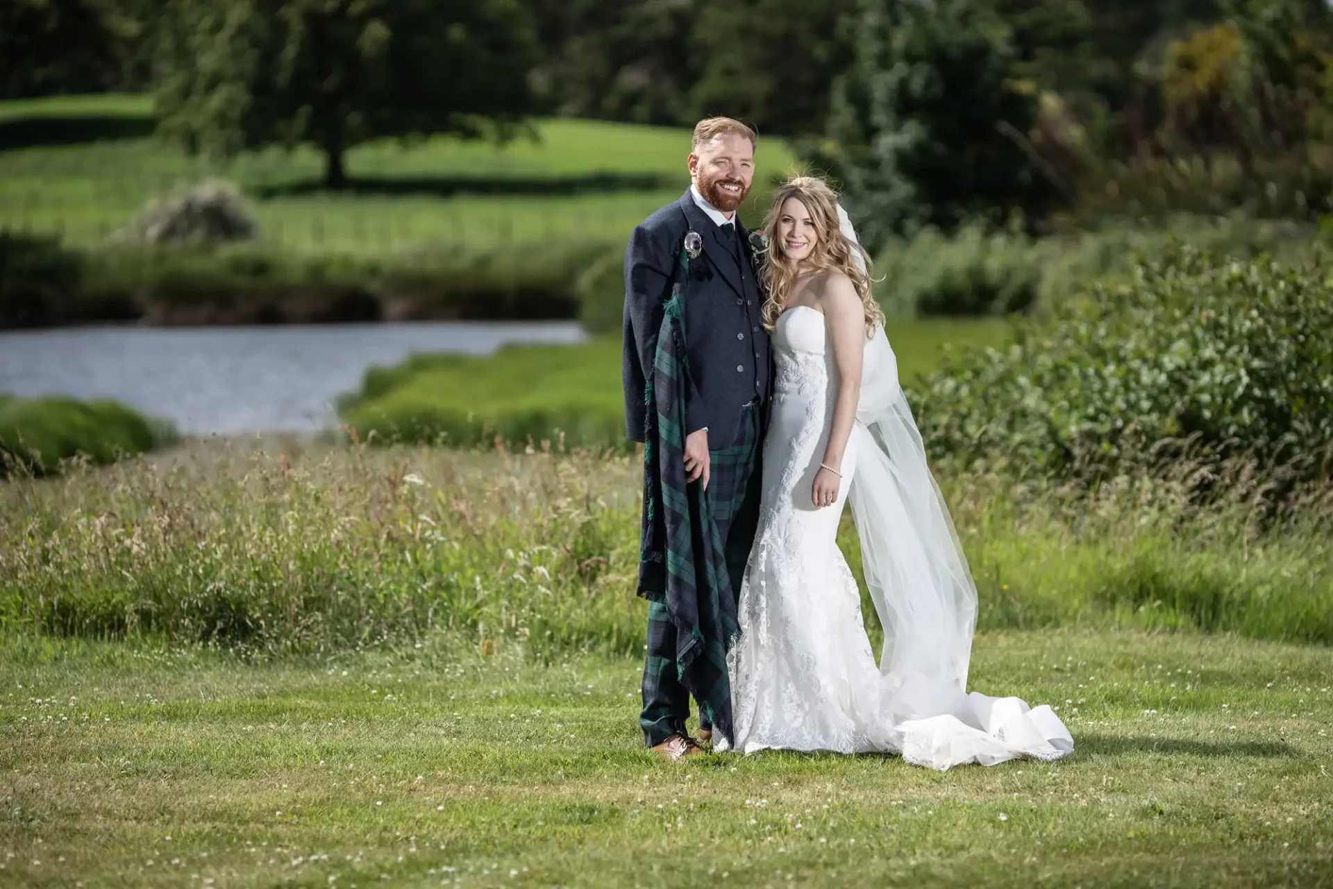 A bride and groom smiling outdoors in formal wedding attire, with a lush green backdrop and a small pond.