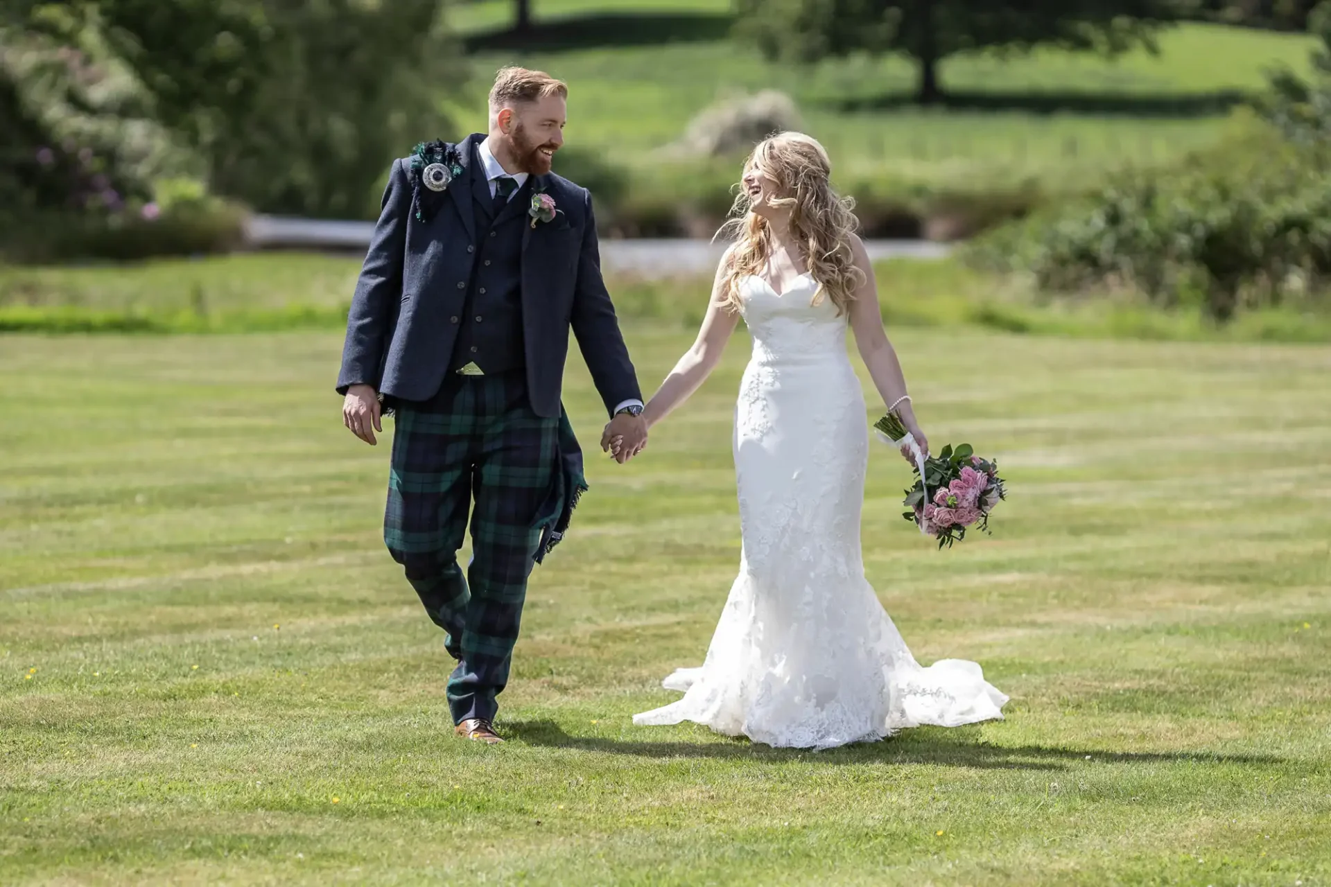 A bride in a white dress and a groom in a tartan kilt walk hand in hand across a grassy field on a sunny day, both smiling.