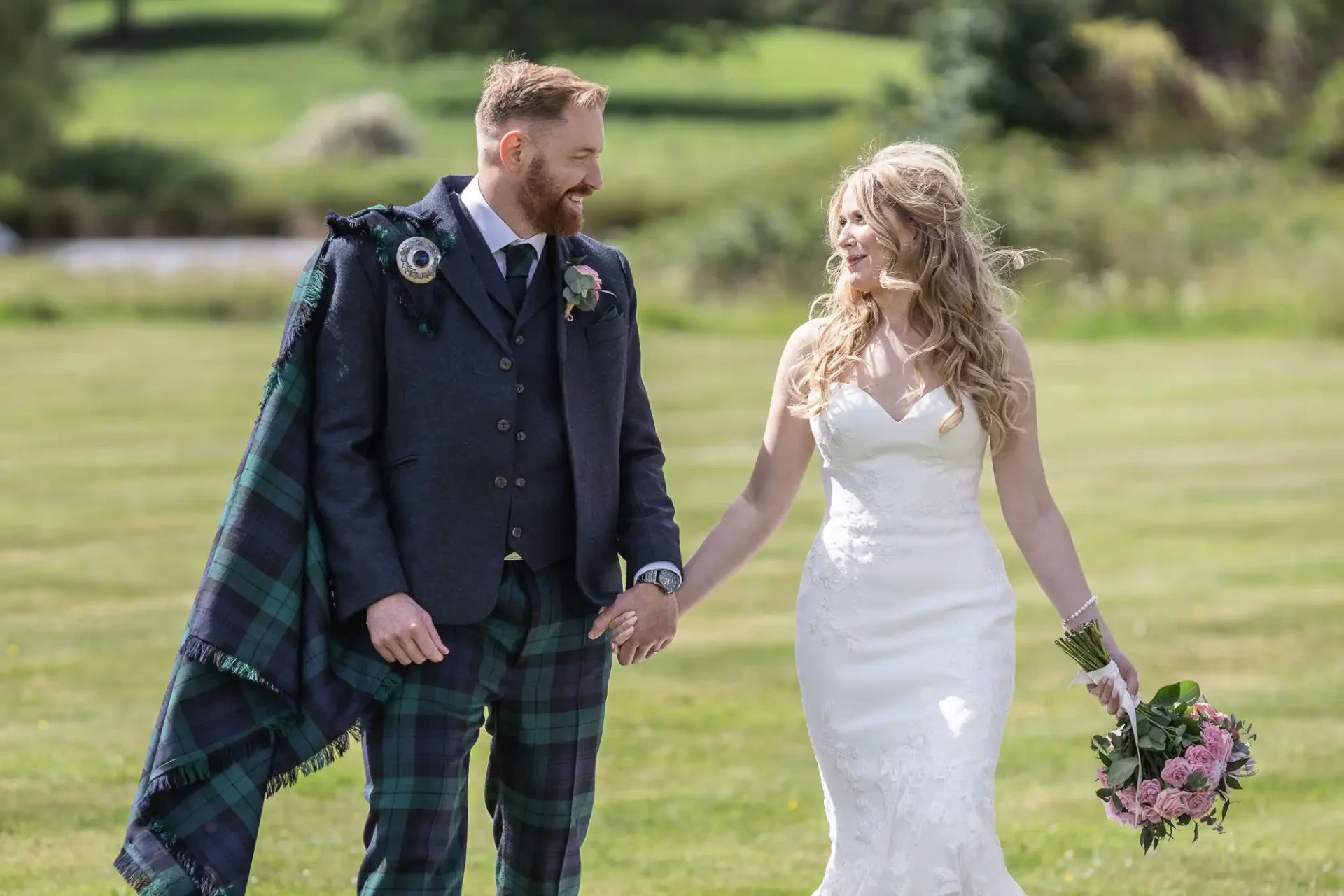 A bride and groom in wedding attire, walking hand-in-hand across a grassy field, the groom in a kilt, both smiling joyfully.