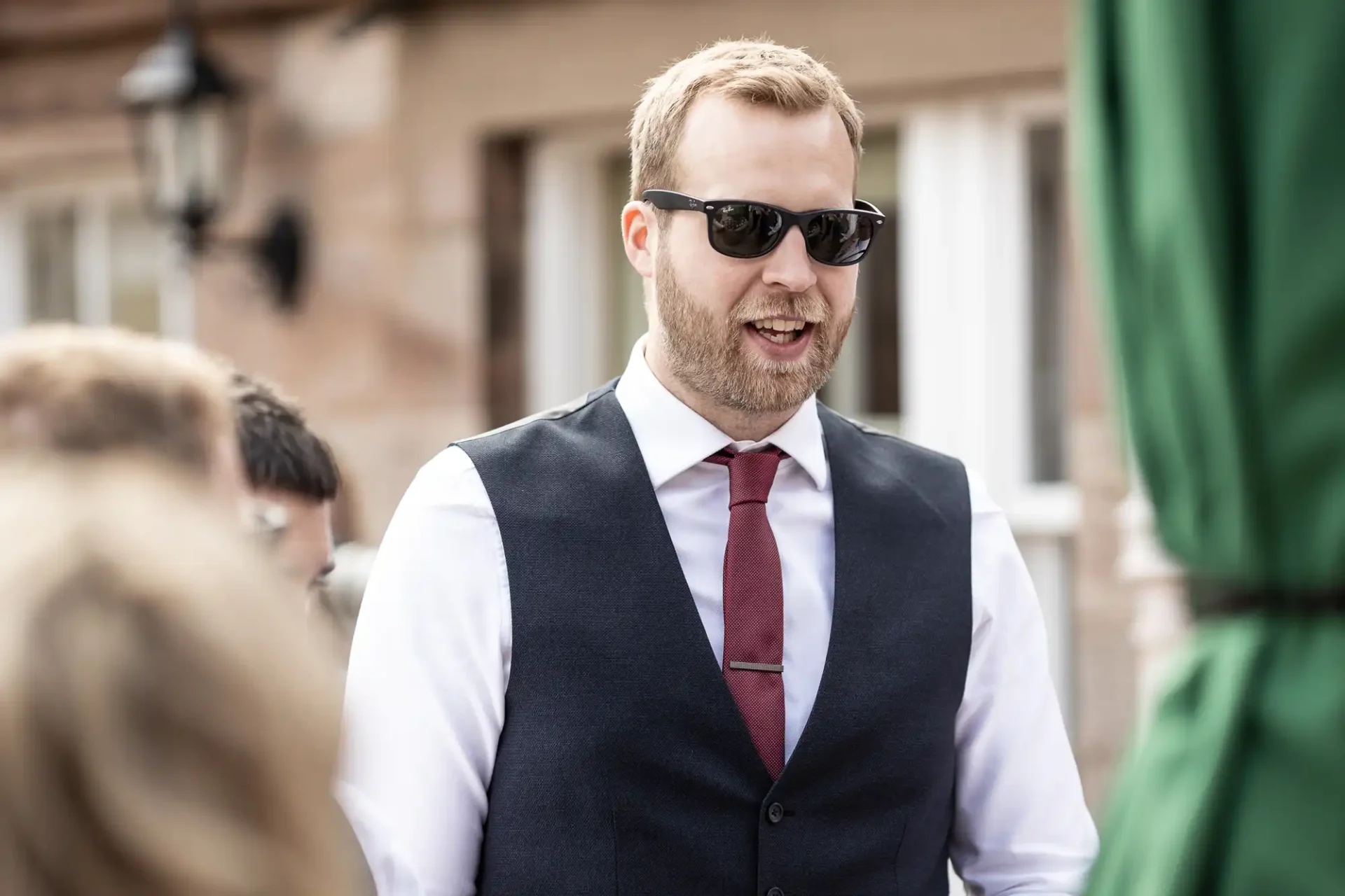 A man in a tailored vest and tie wearing sunglasses talks to people outdoors.