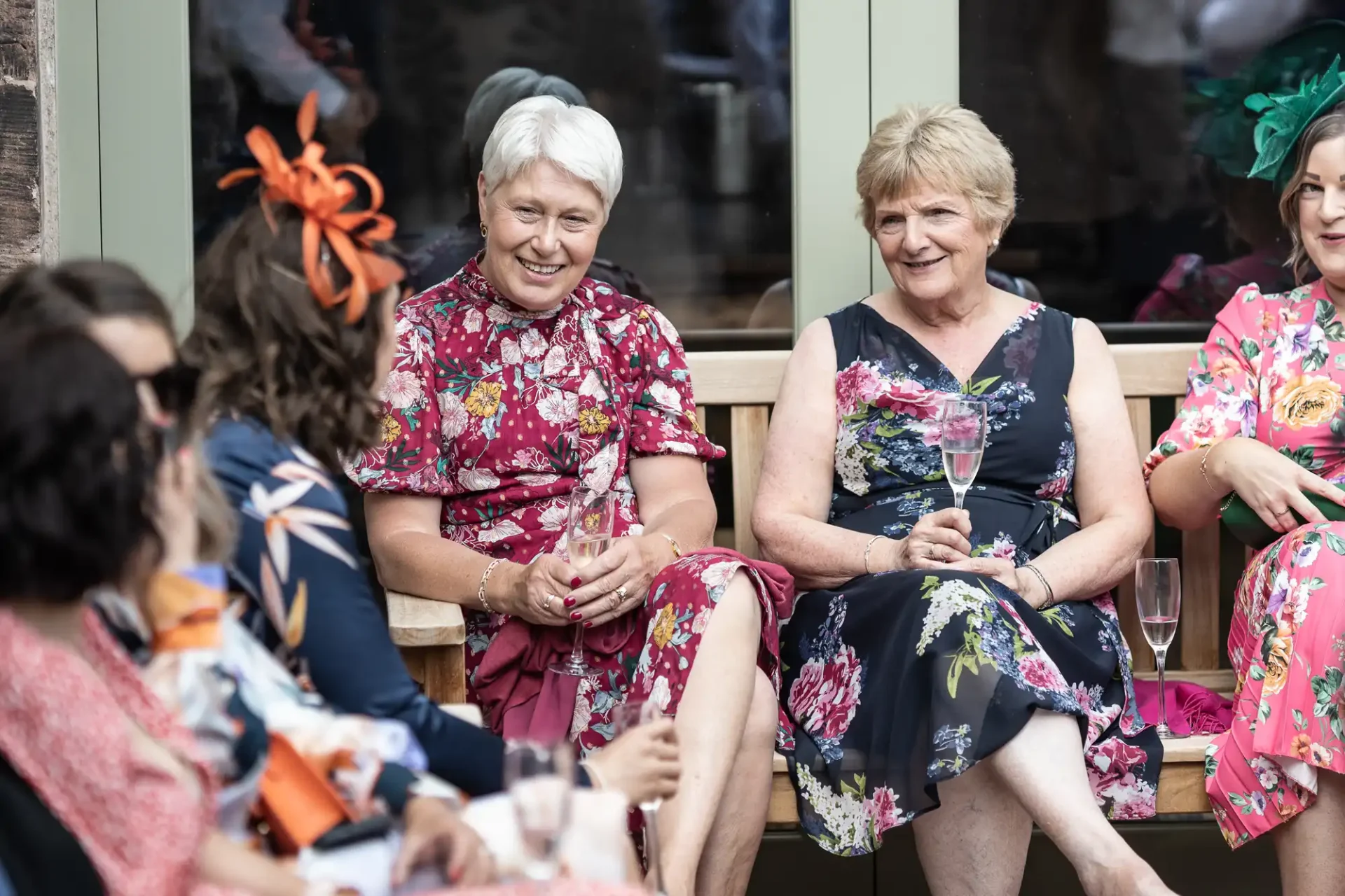 Four women in floral dresses with hats or fascinators sitting and smiling, one holding a champagne flute, at an outdoor social event.