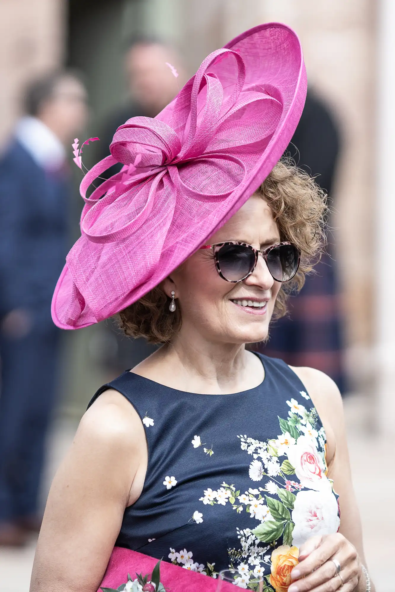 A woman with curly hair wearing a large pink hat and a floral dress smiles, holding a rose, outdoors at a formal event.