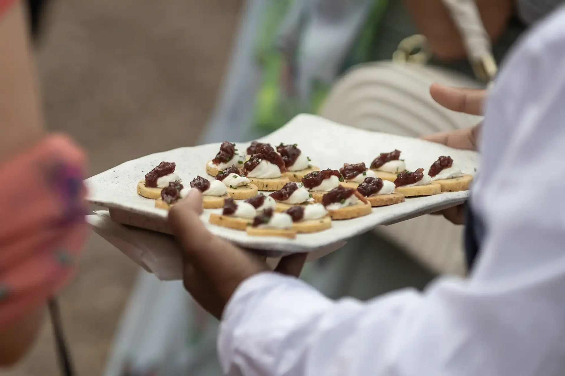A person holds a tray of hors d'oeuvres, featuring small toasts topped with cheese and a dark fruit compote, at an outdoor event.