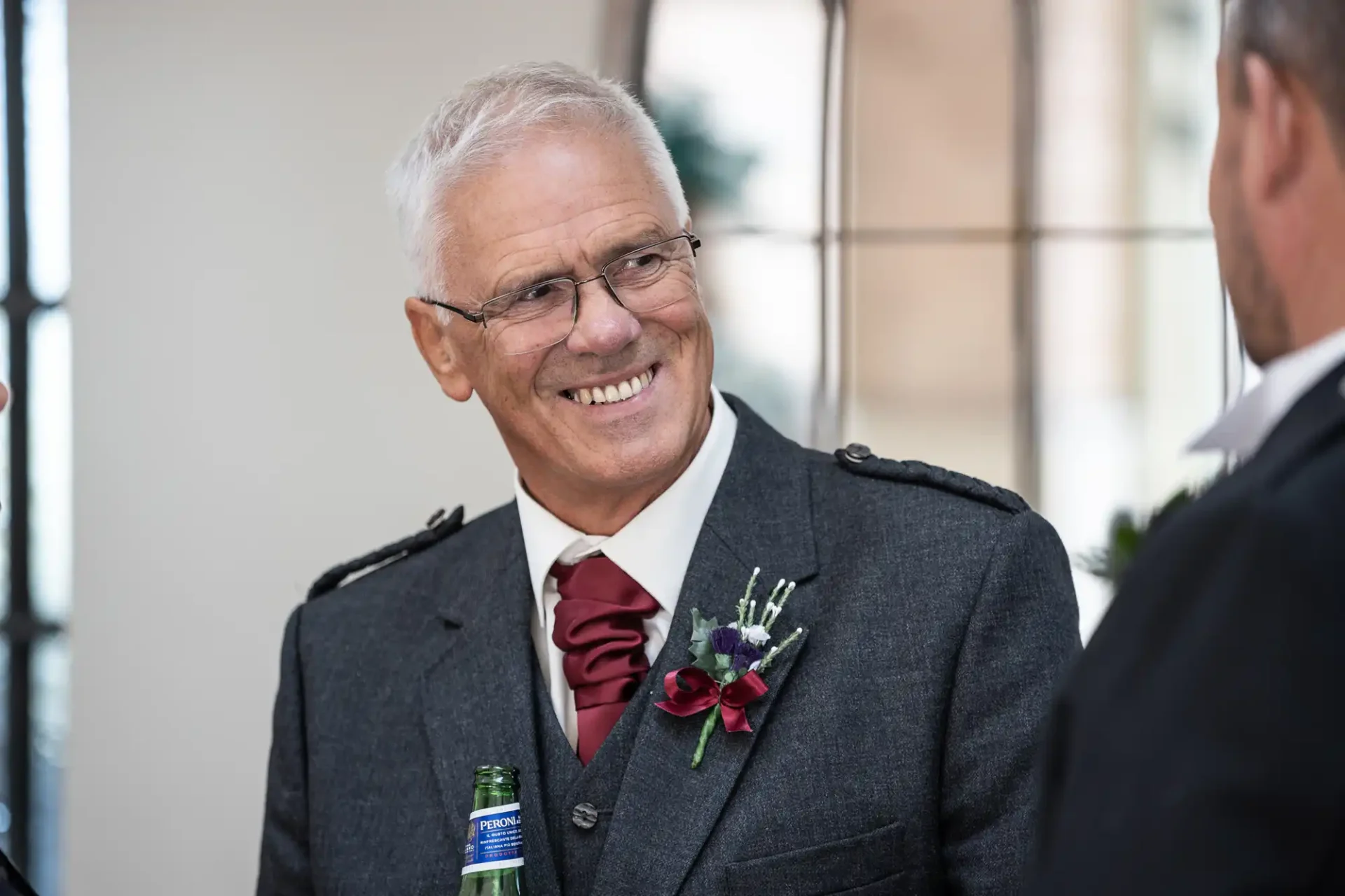 An older man in a gray suit, smiling, with a red flower boutonniere, holding a green bottle, talking to another person out of view.