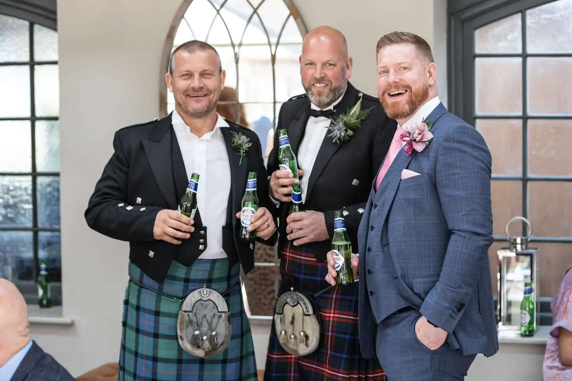 Three smiling men in formal attire with tartan kilts, holding beer bottles at a social event.