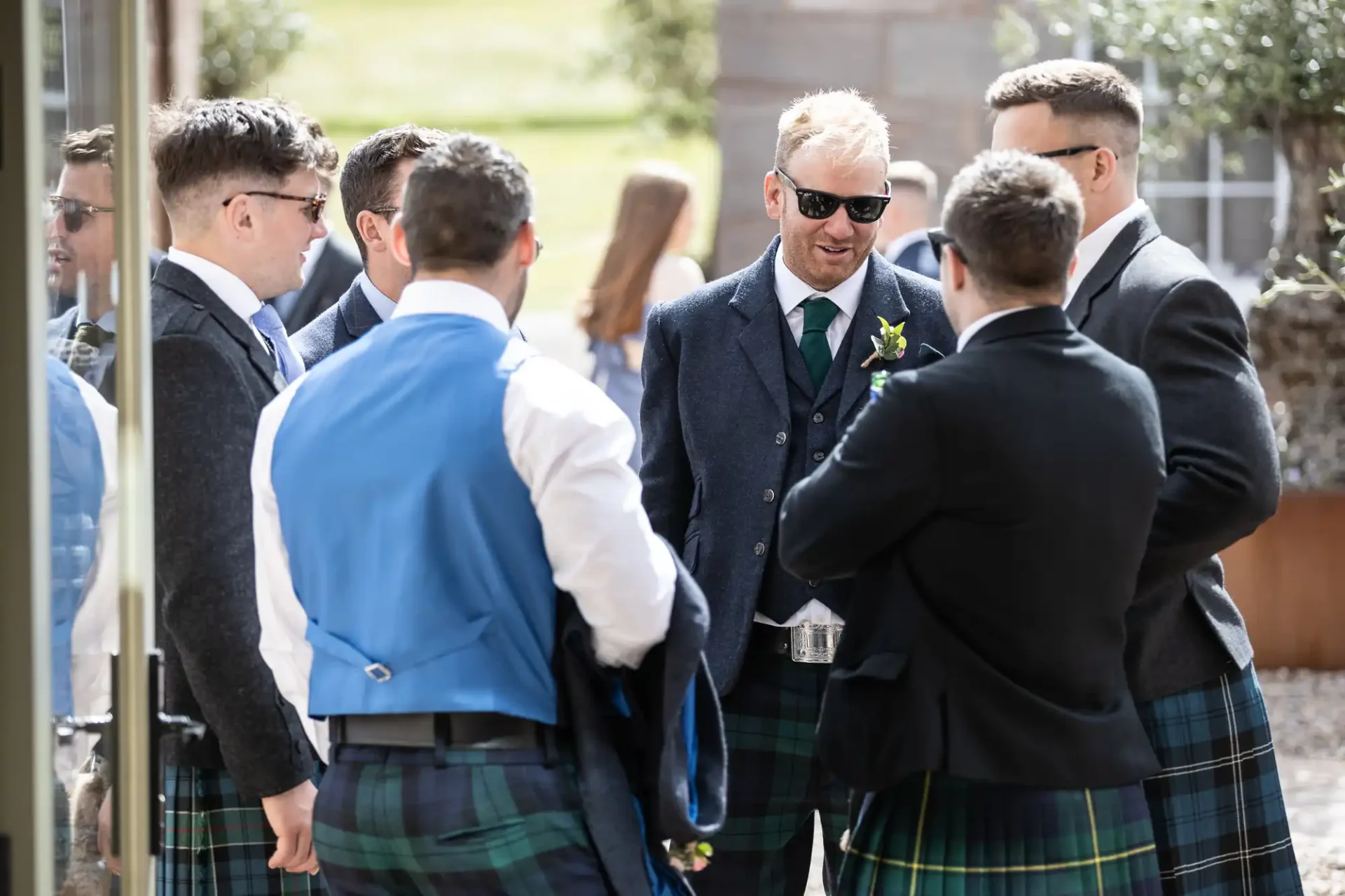 A group of men in kilts and suits conversing outdoors at a formal event, with one man smiling towards the camera.