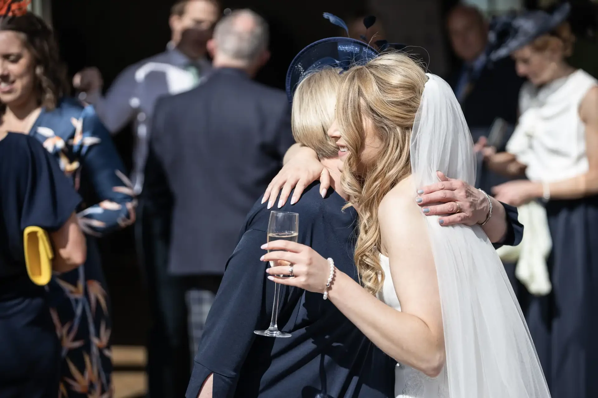 A bride in a white dress and veil embracing a woman in a navy dress, both smiling, at a sunny outdoor wedding ceremony.