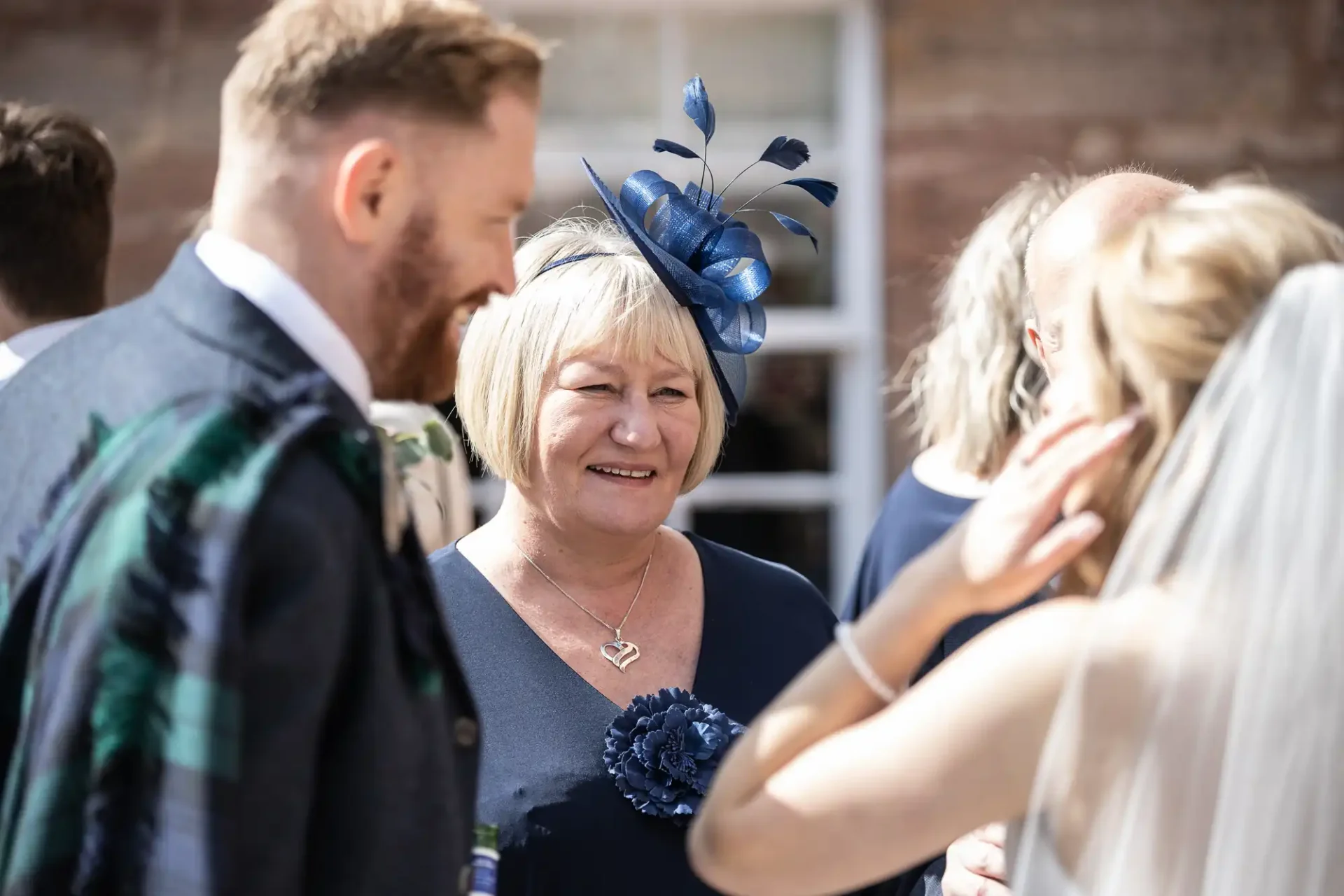 Smiling woman in a blue hat and floral dress at a sunny wedding reception, talking with guests including a man in a tartan waistcoat.