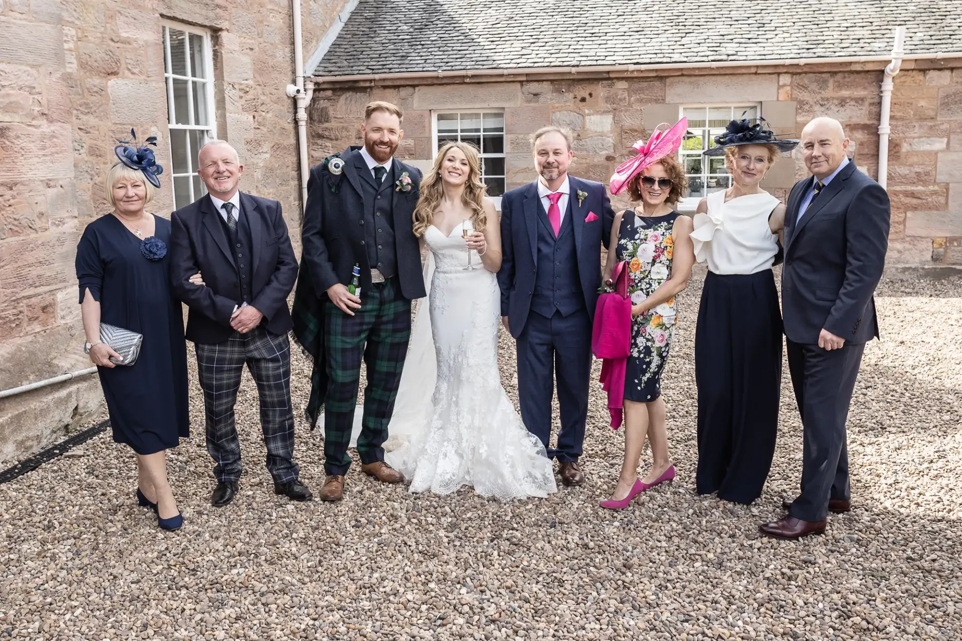 A bride and groom smiling with six guests in formal attire, including kilts and hats, standing in front of a stone building.