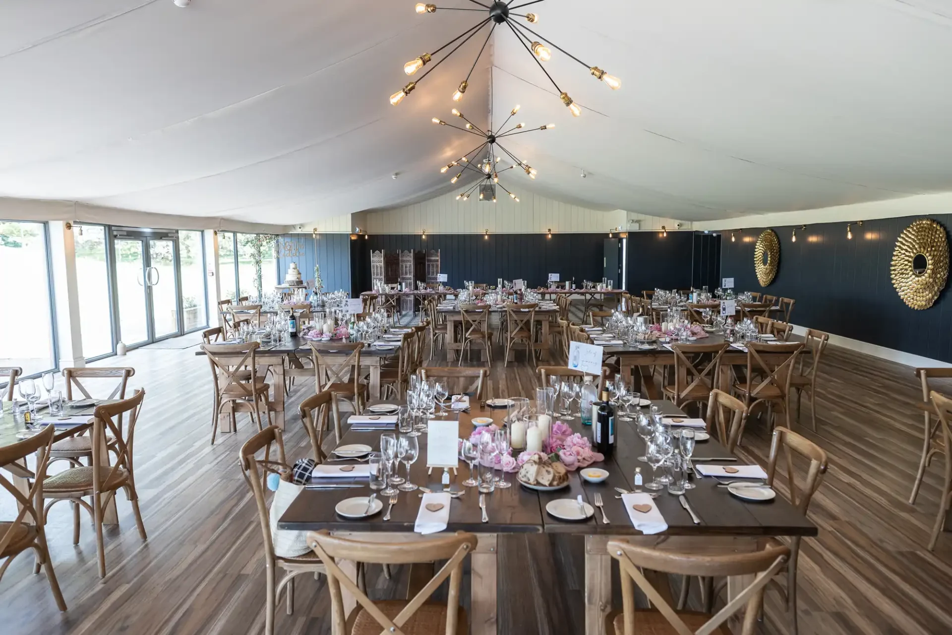 Elegant event venue interior with tables set for a formal dining, featuring wooden chairs and floral centerpieces under a tent with string lights.