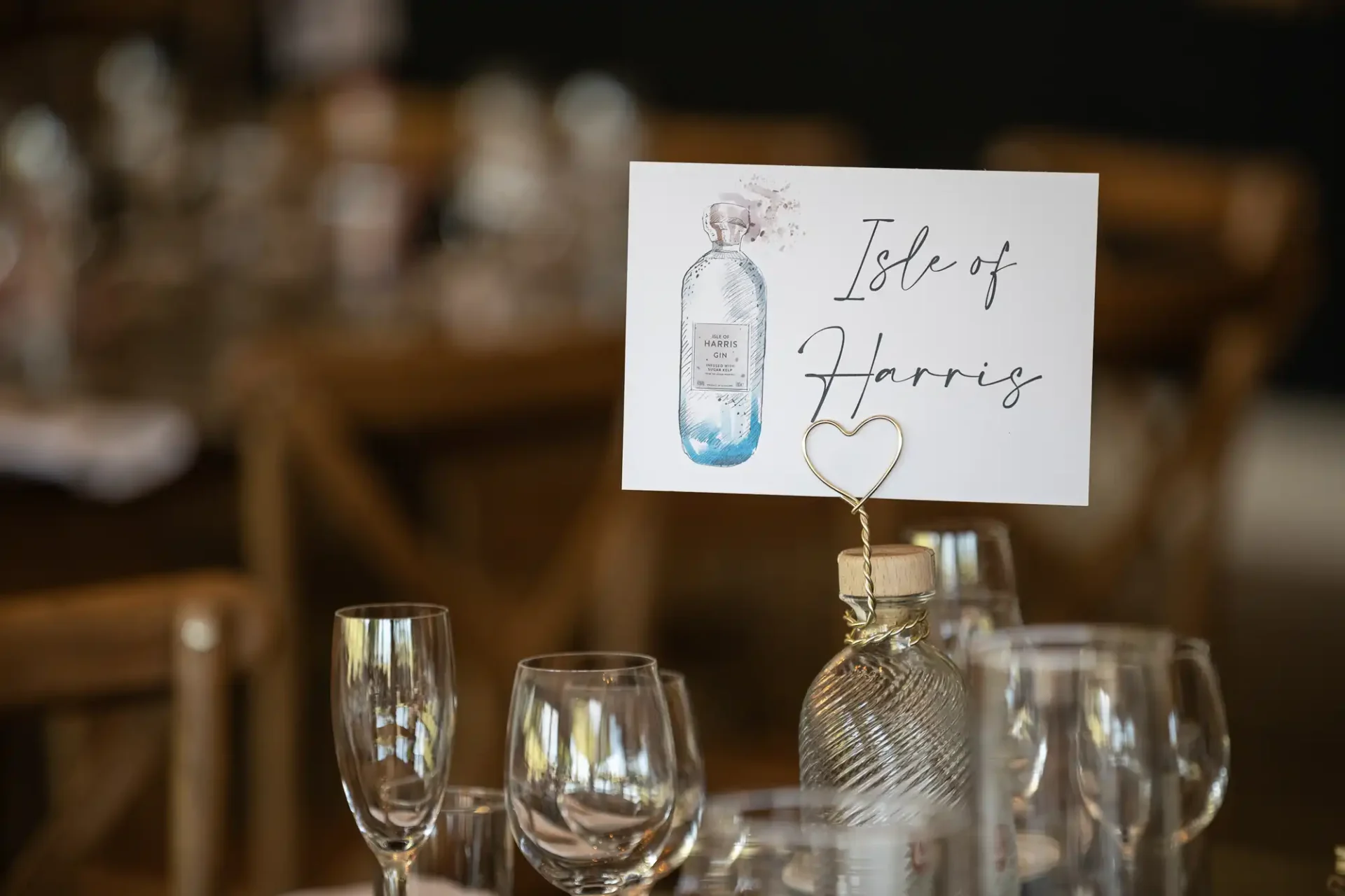 A table setting at an event with a name card reading "isle of harris" propped on a heart-shaped holder, flanked by wine glasses and blurred background.