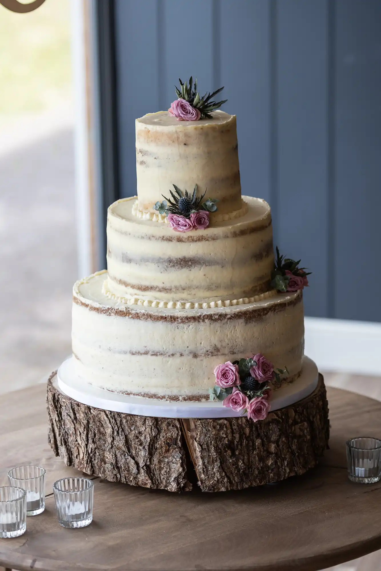 Three-tiered wedding cake with white icing and pink floral decorations on a wooden stand, placed next to clear glasses on a table.
