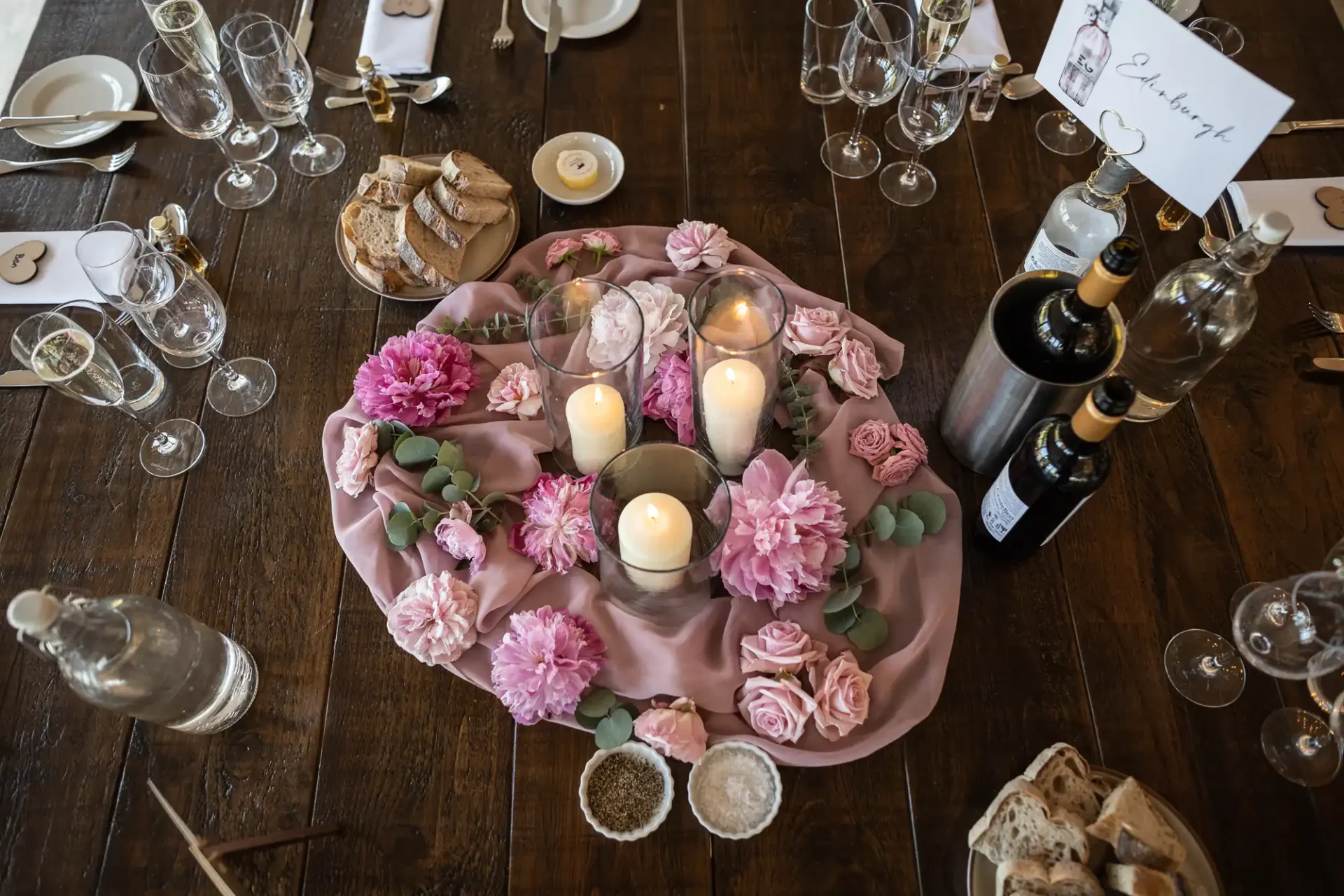 Elegant dining table setup with pink floral centerpieces, candles, wine bottles, and filled glasses on a rustic wooden surface.