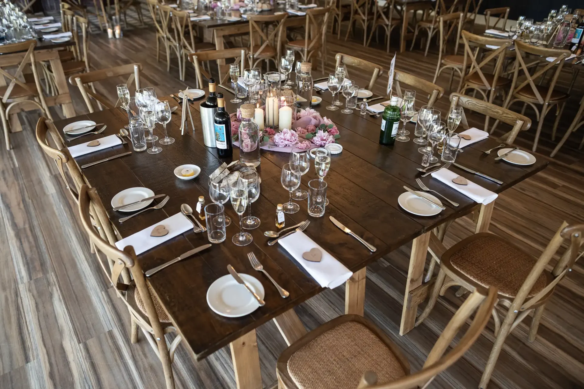 Elegantly set tables at a banquet hall with wooden chairs, floral centerpieces, candles, and dinnerware.