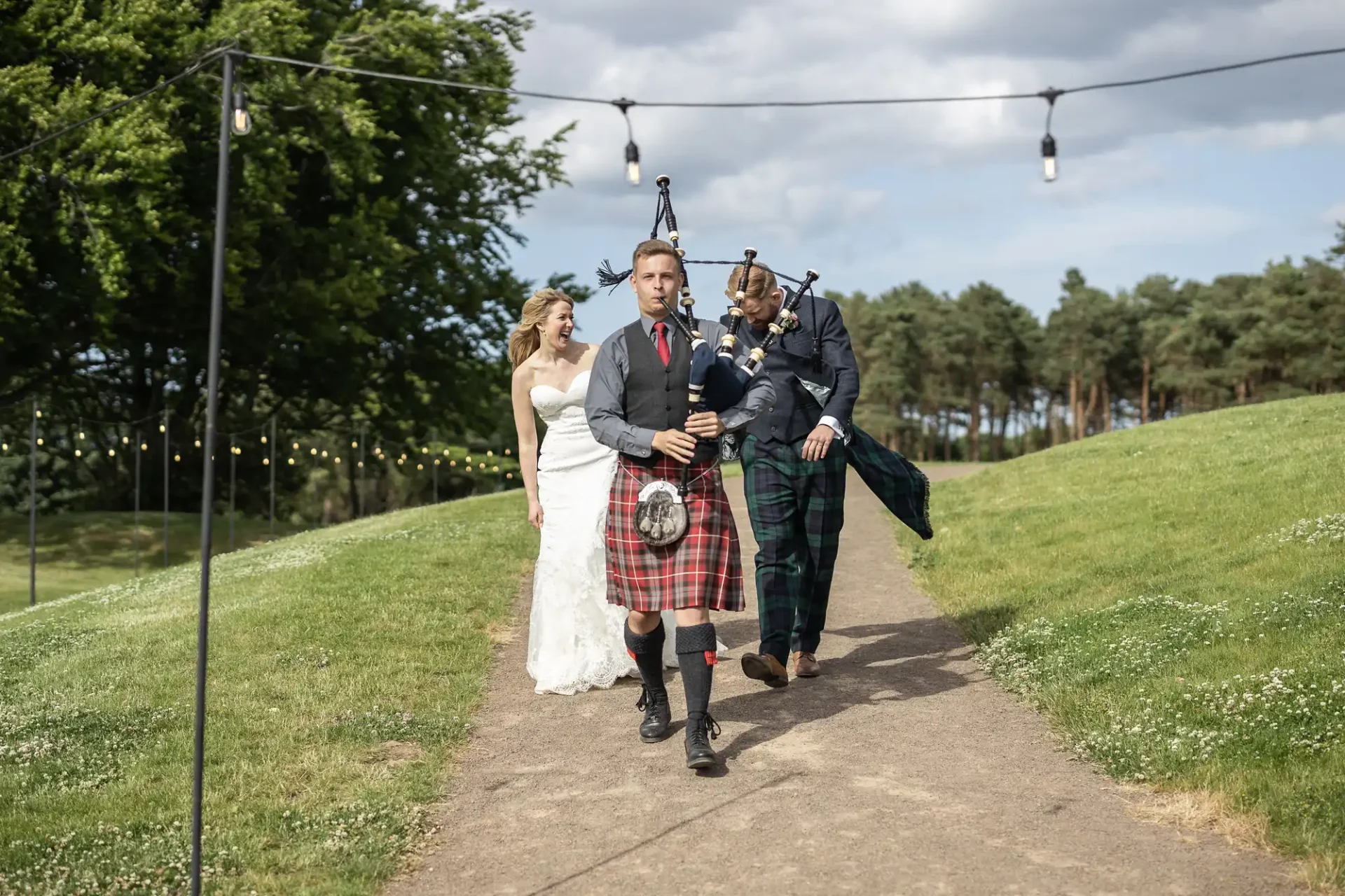 A bride and groom walk along a pathway with a bagpiper in traditional scottish attire, outdoor setting with greenery and string lights overhead.