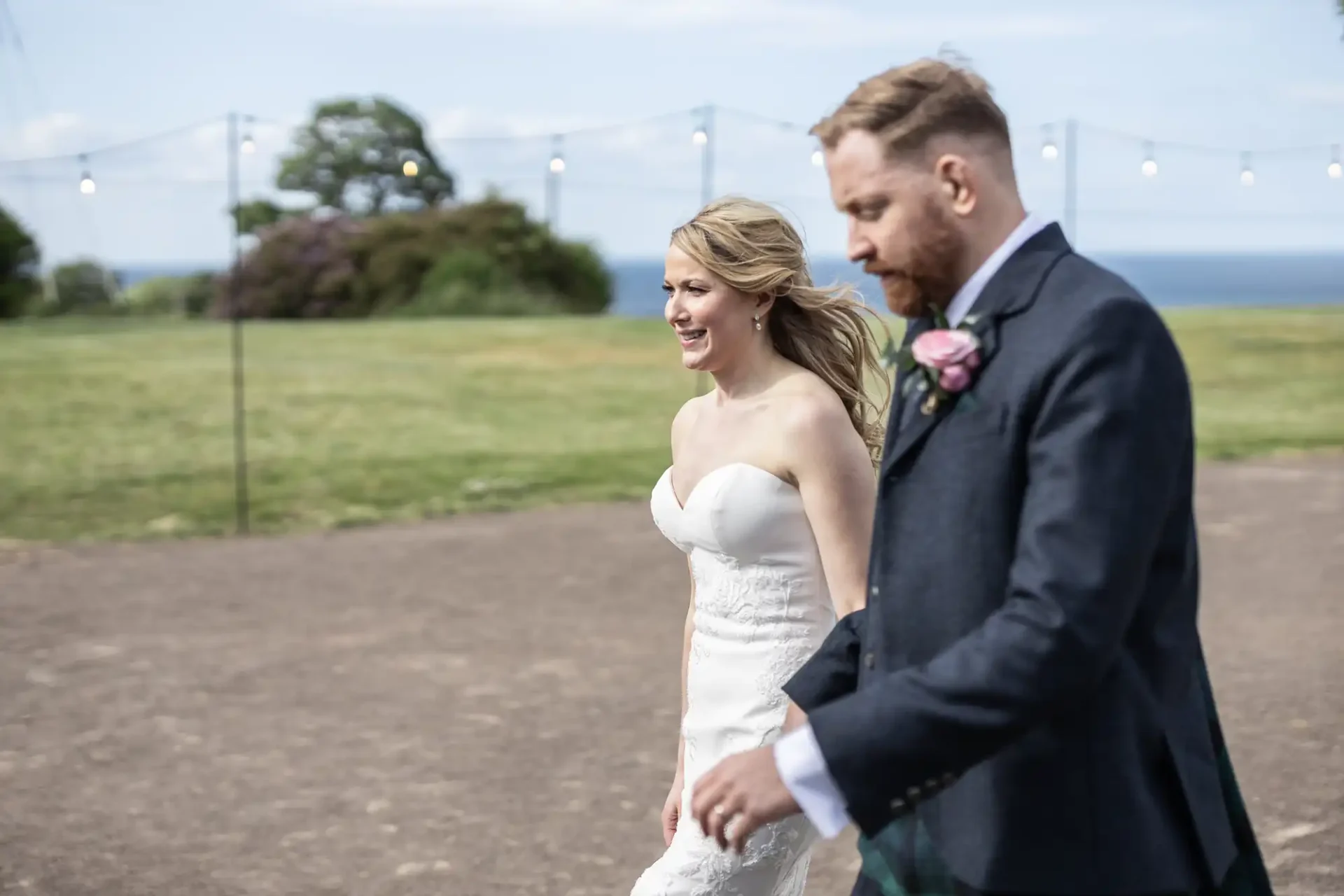 A bride and groom walking hand in hand outdoors, both smiling, on a sunny day with string lights and trees in the background.